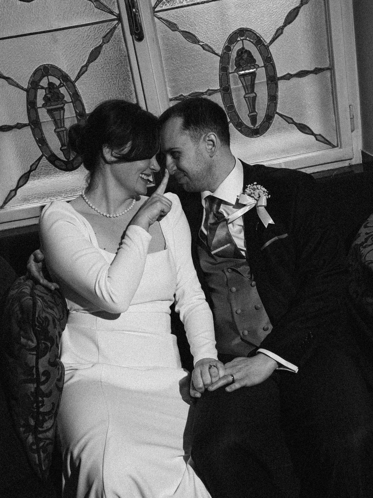 Black and white photo of a couple on their wedding day, smiling and touching noses, with the bride wearing a wedding dress and pearl necklace and the groom wearing a suit with a boutonniere, holding hands.