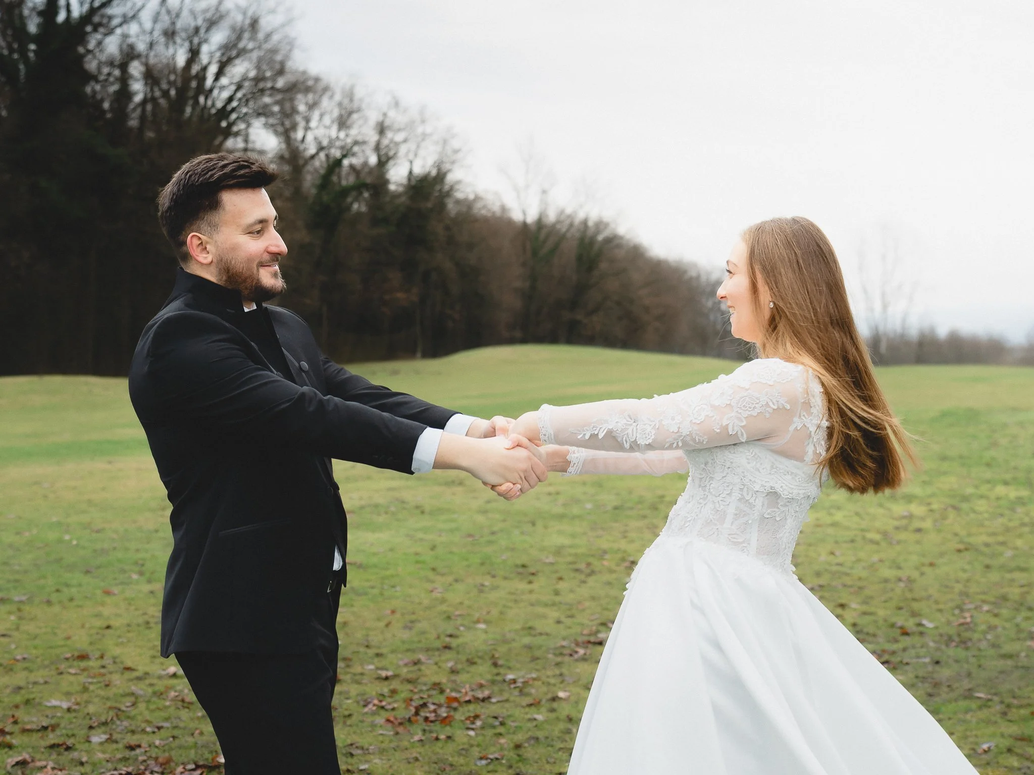 A bride and groom holding hands and smiling at each other outdoors in a grassy field with trees in the background.