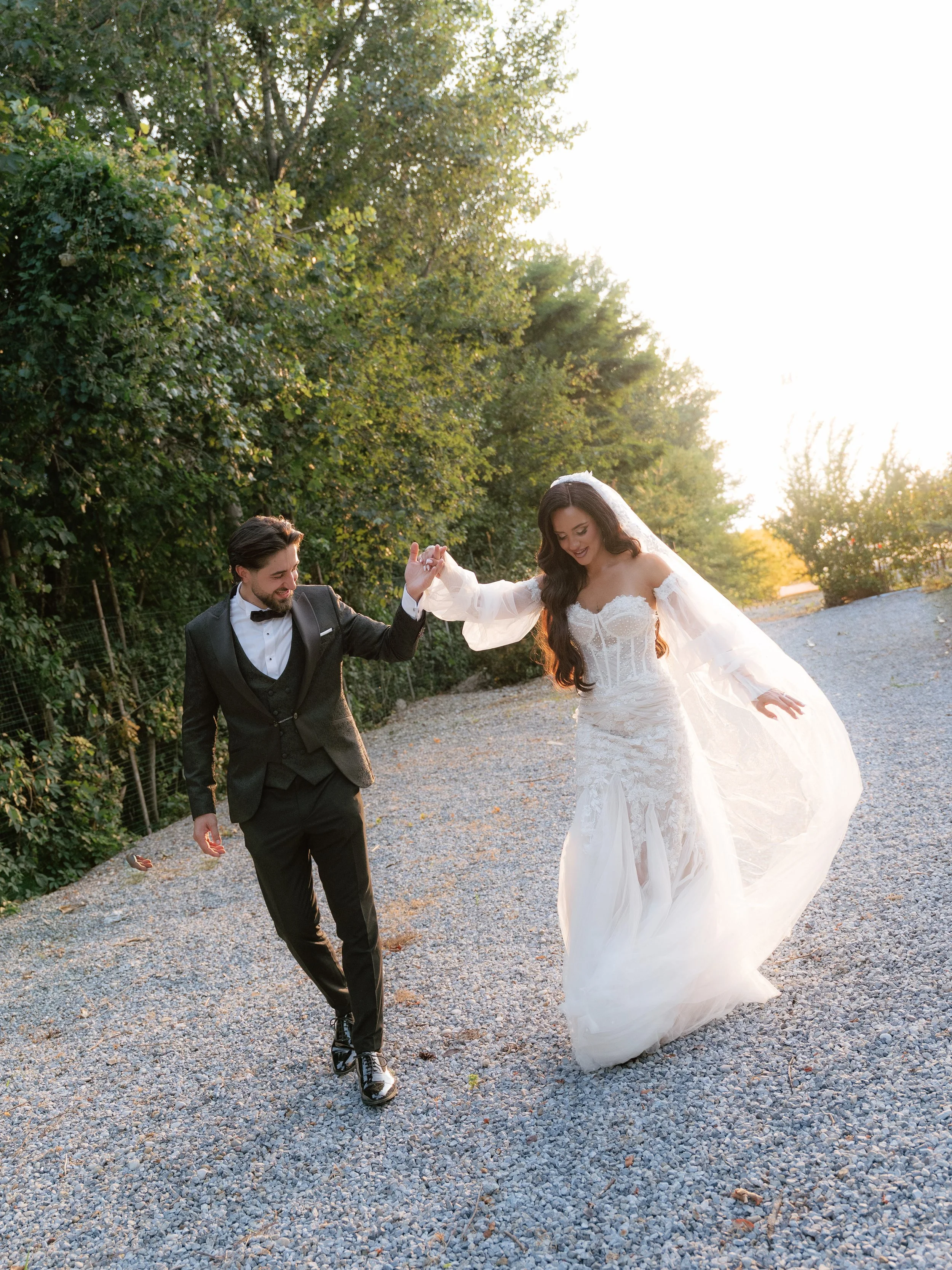 A bride and groom dancing outdoors on a gravel path surrounded by trees during sunset, holding hands and smiling.