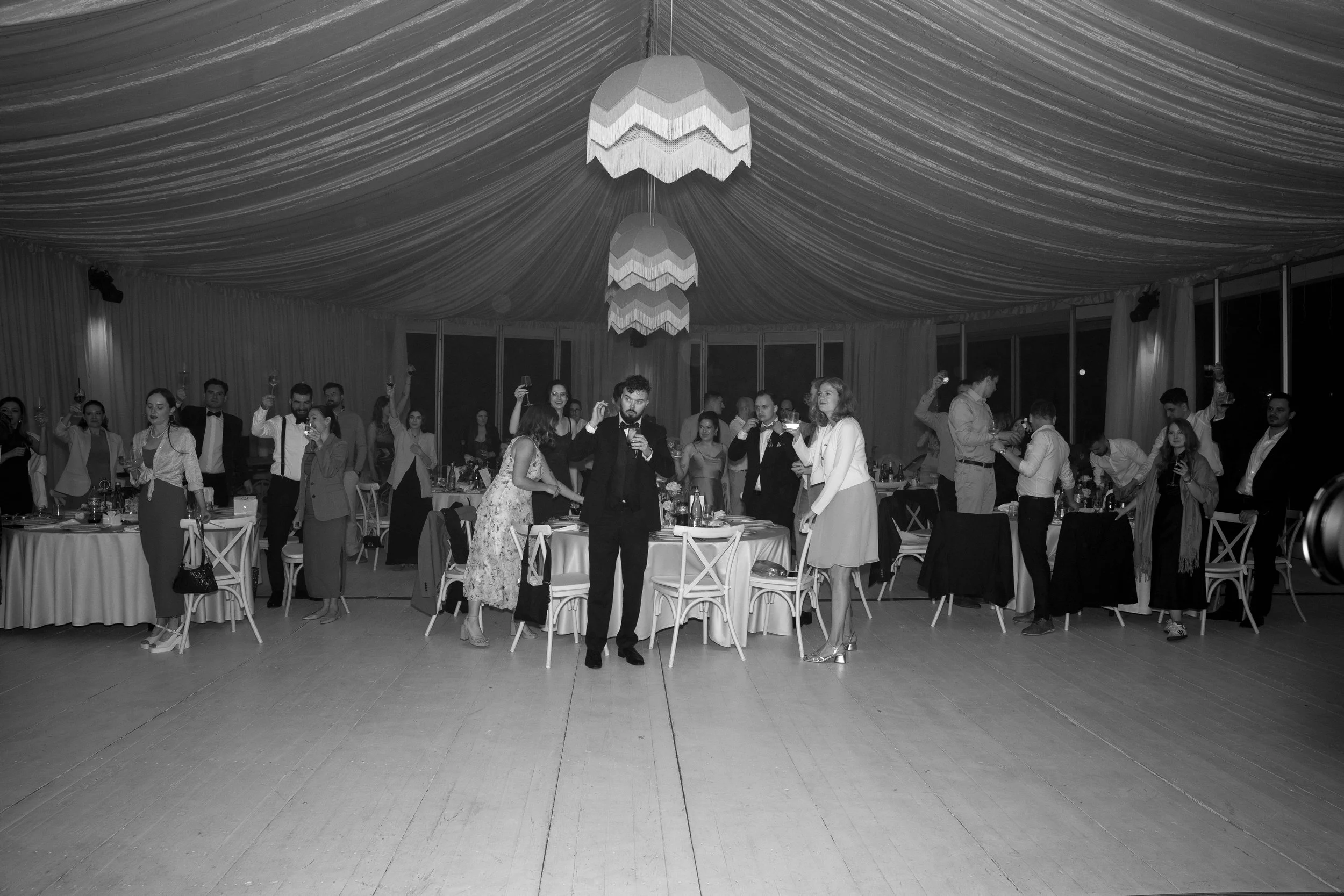 A group of people at a formal event or party inside a decorated banquet hall, raising glasses in a celebratory toast, with tables and chairs and hanging ceiling lights.