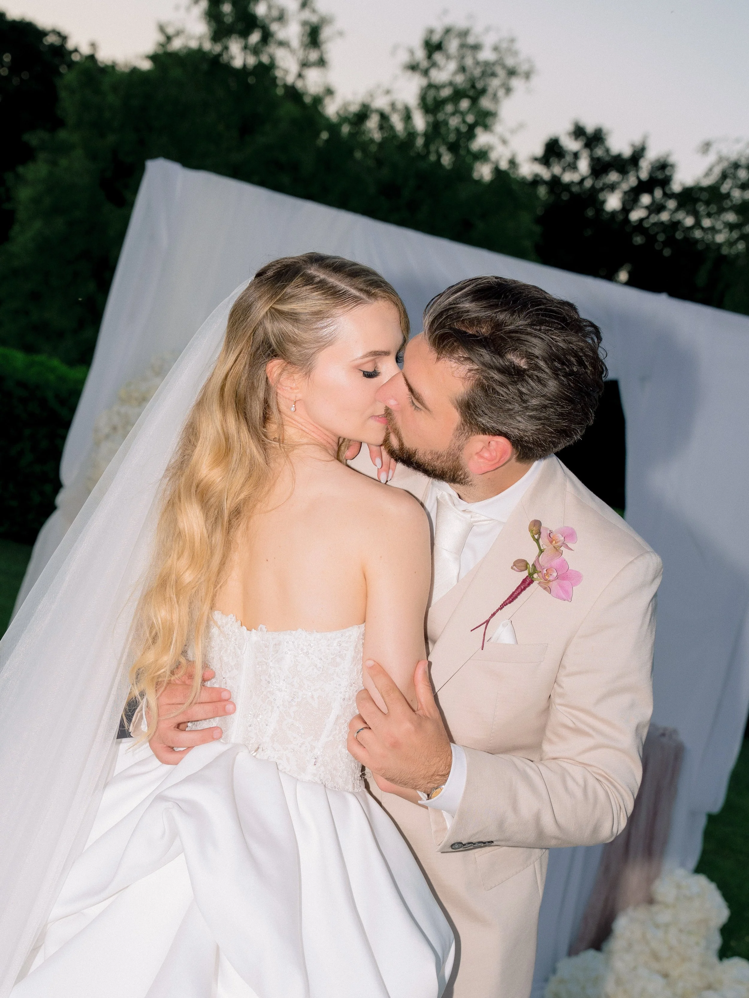 A bride and groom kiss during their wedding ceremony outdoors, with the bride wearing a white wedding gown and veil, and the groom in a beige suit with a pink orchid boutonniere.