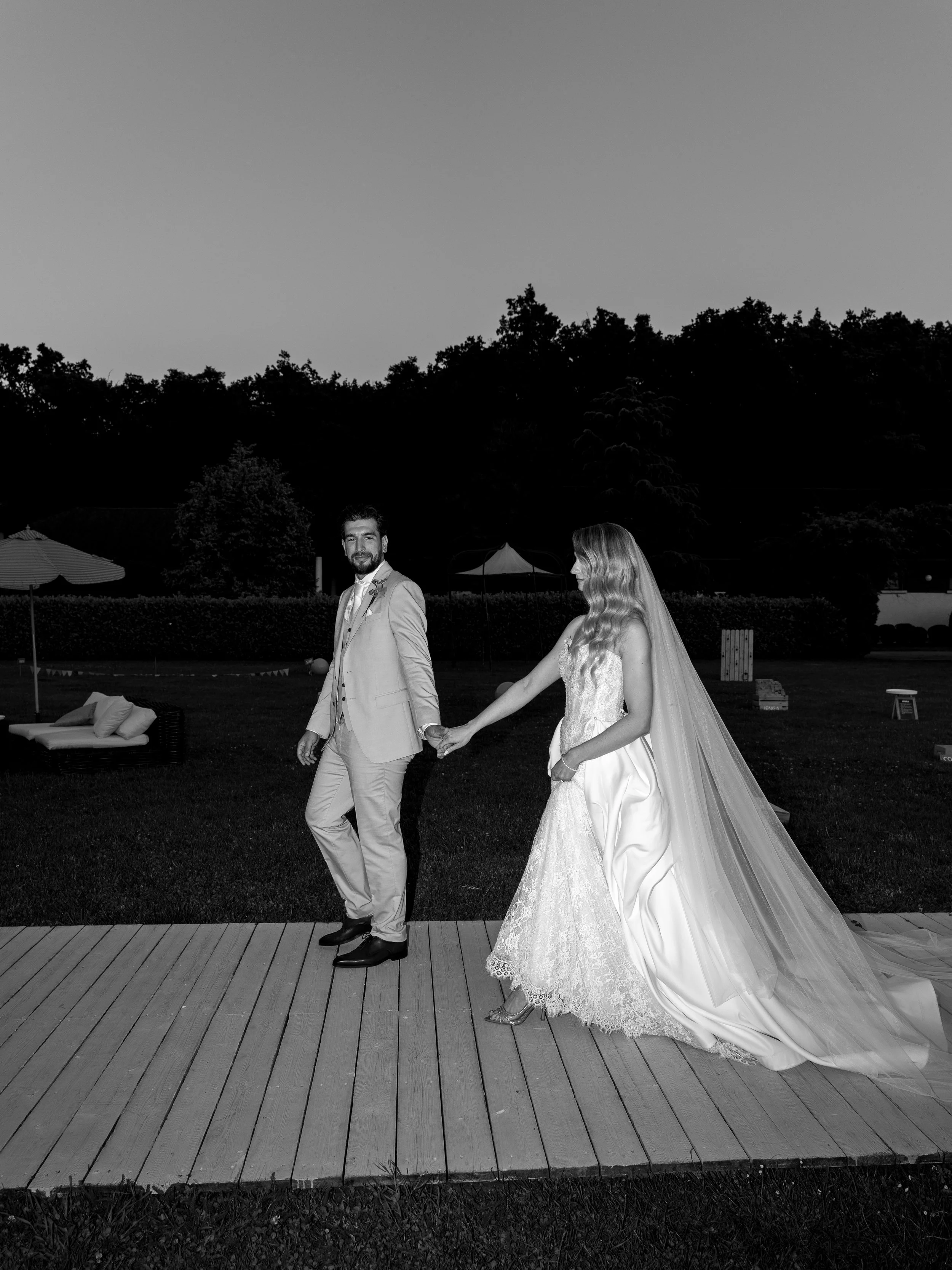 A bride and groom holding hands outdoors during their wedding at dusk, standing on a wooden deck with trees and outdoor furniture in the background, black and white photo.