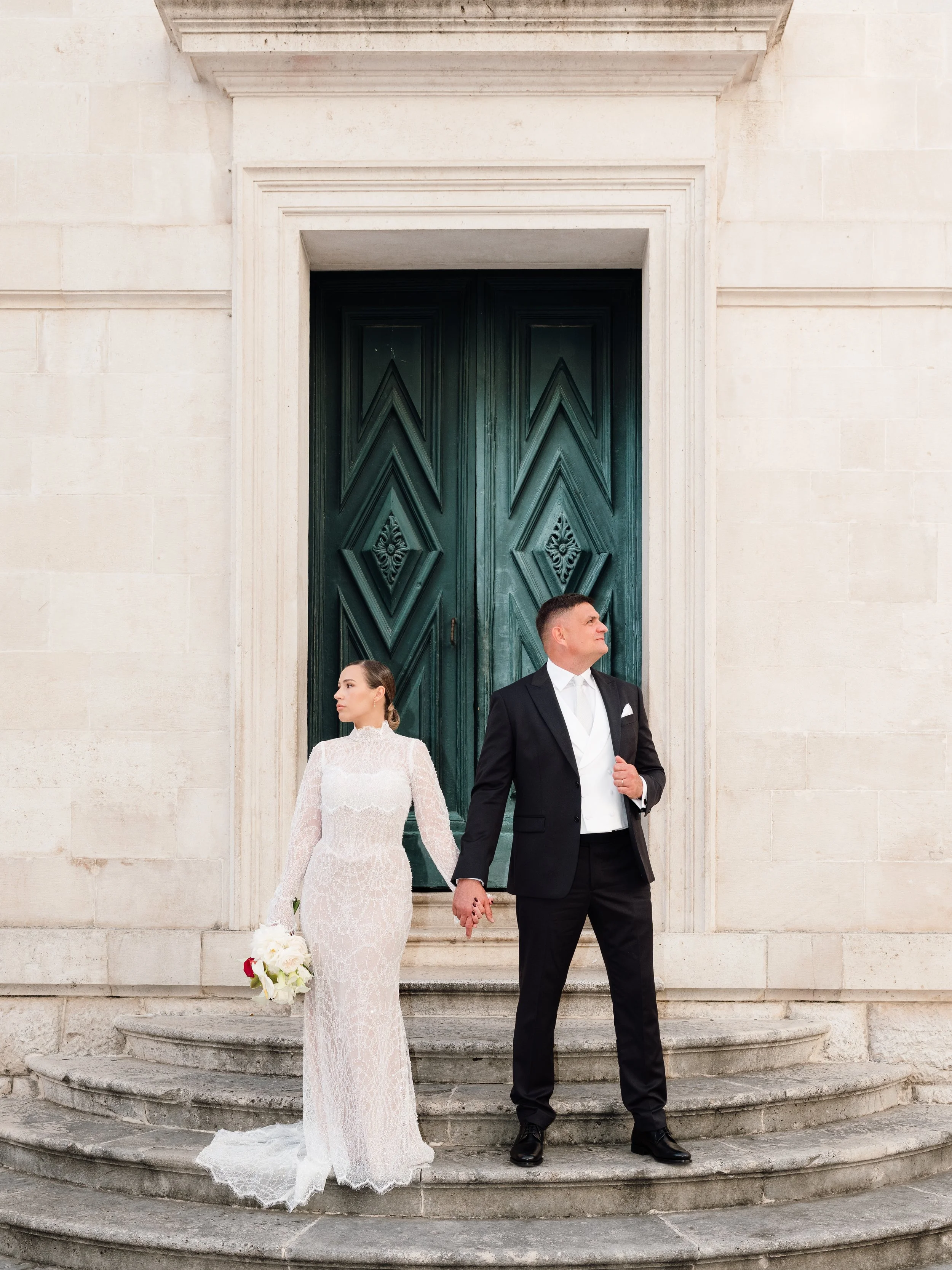 A bride and groom standing on stone steps in front of a large, ornate green door, holding hands, with the bride holding a bouquet of white flowers.
