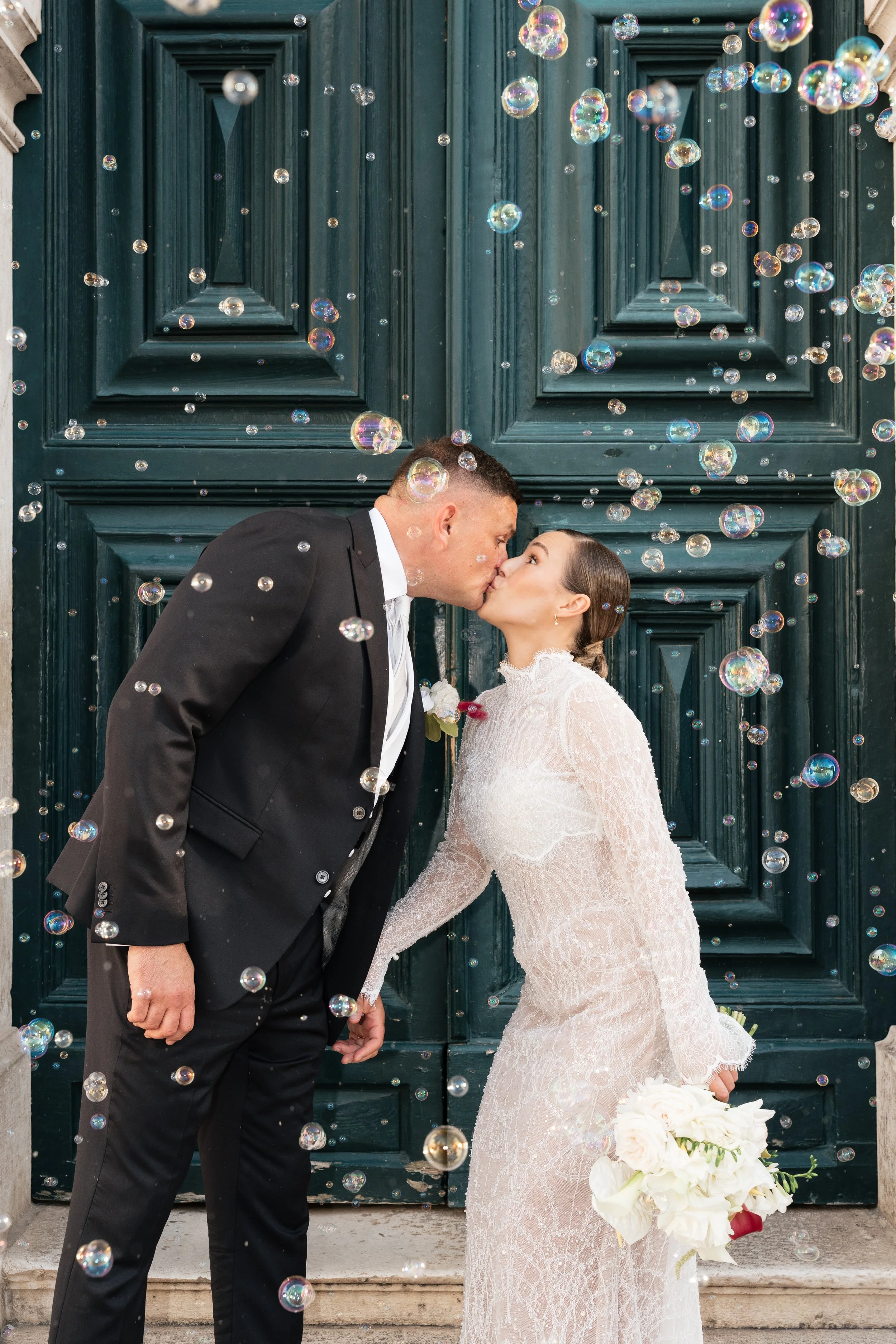 A bride and groom kissing in front of a large green wooden door, surrounded by floating bubbles. The bride is holding a bouquet of white flowers.