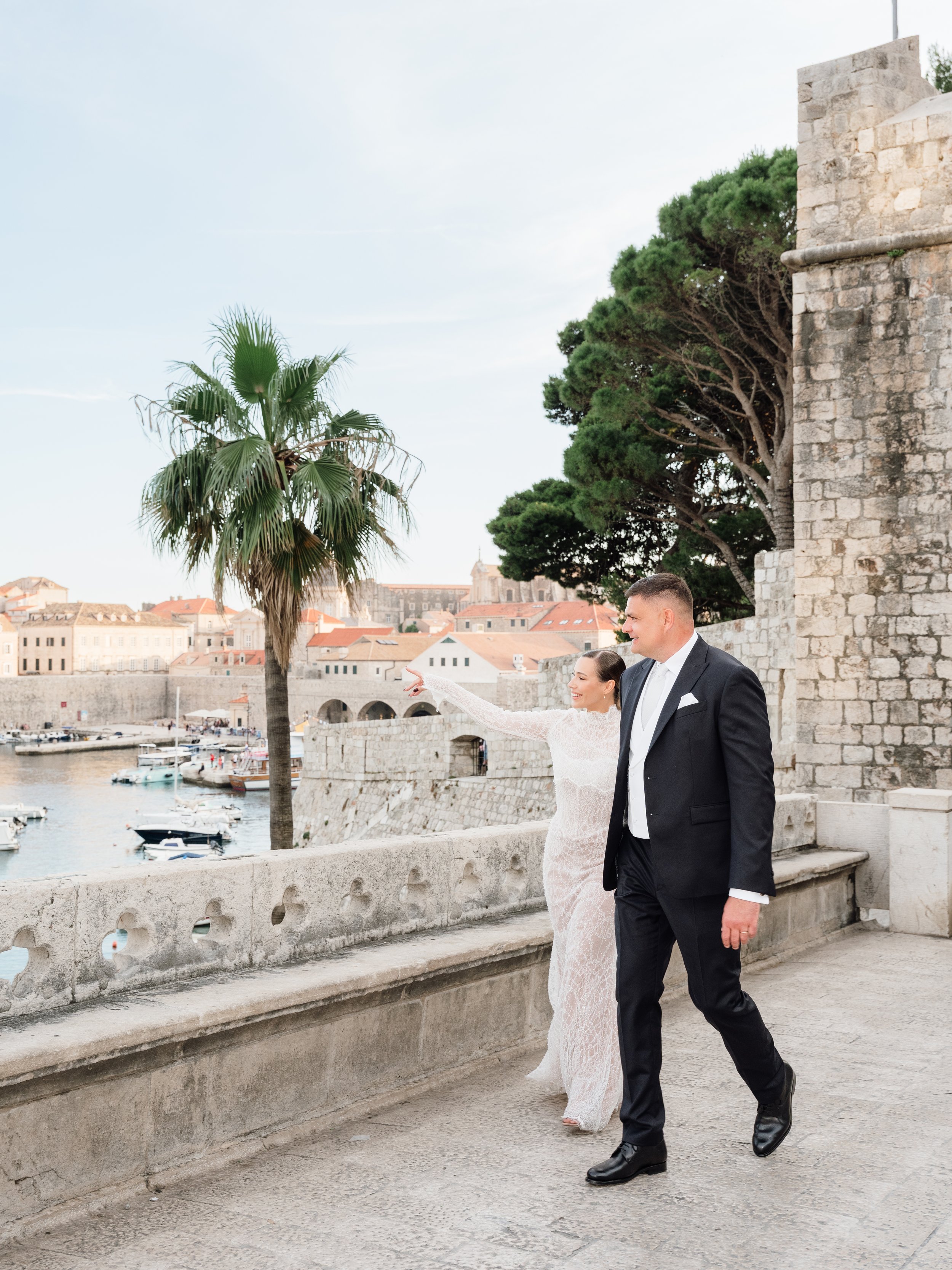 A bride and groom walking along a stone railing by the water in a historic city. The bride is pointing towards something in the distance, and they are smiling. The background features a large palm tree, historic buildings, and boats in the water.