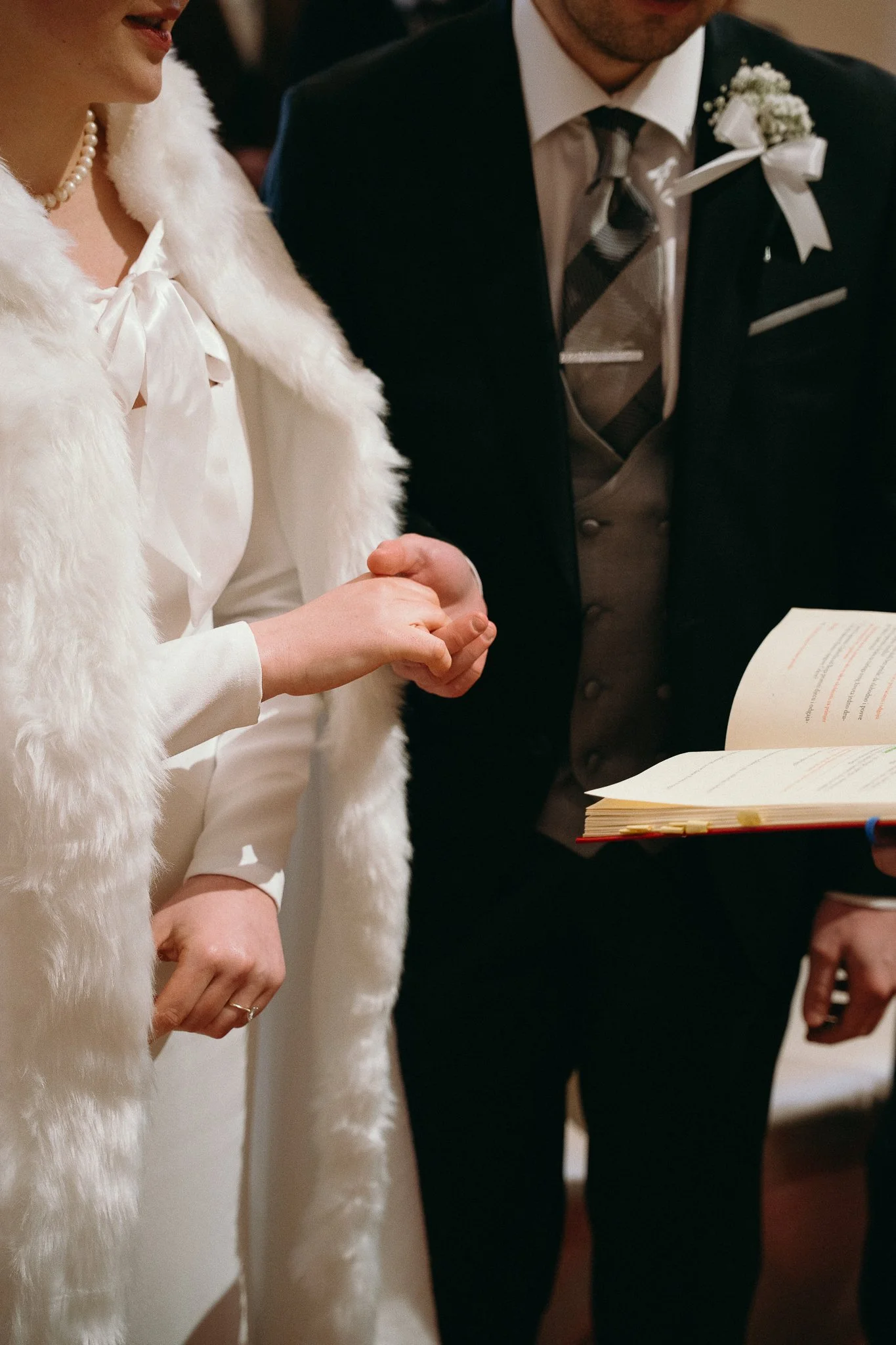 Close-up of a wedding ceremony with the bride and groom holding hands, the bride wearing a white dress with a faux fur shawl, and the groom in a dark suit with a floral boutonniere, reading from a wedding book.