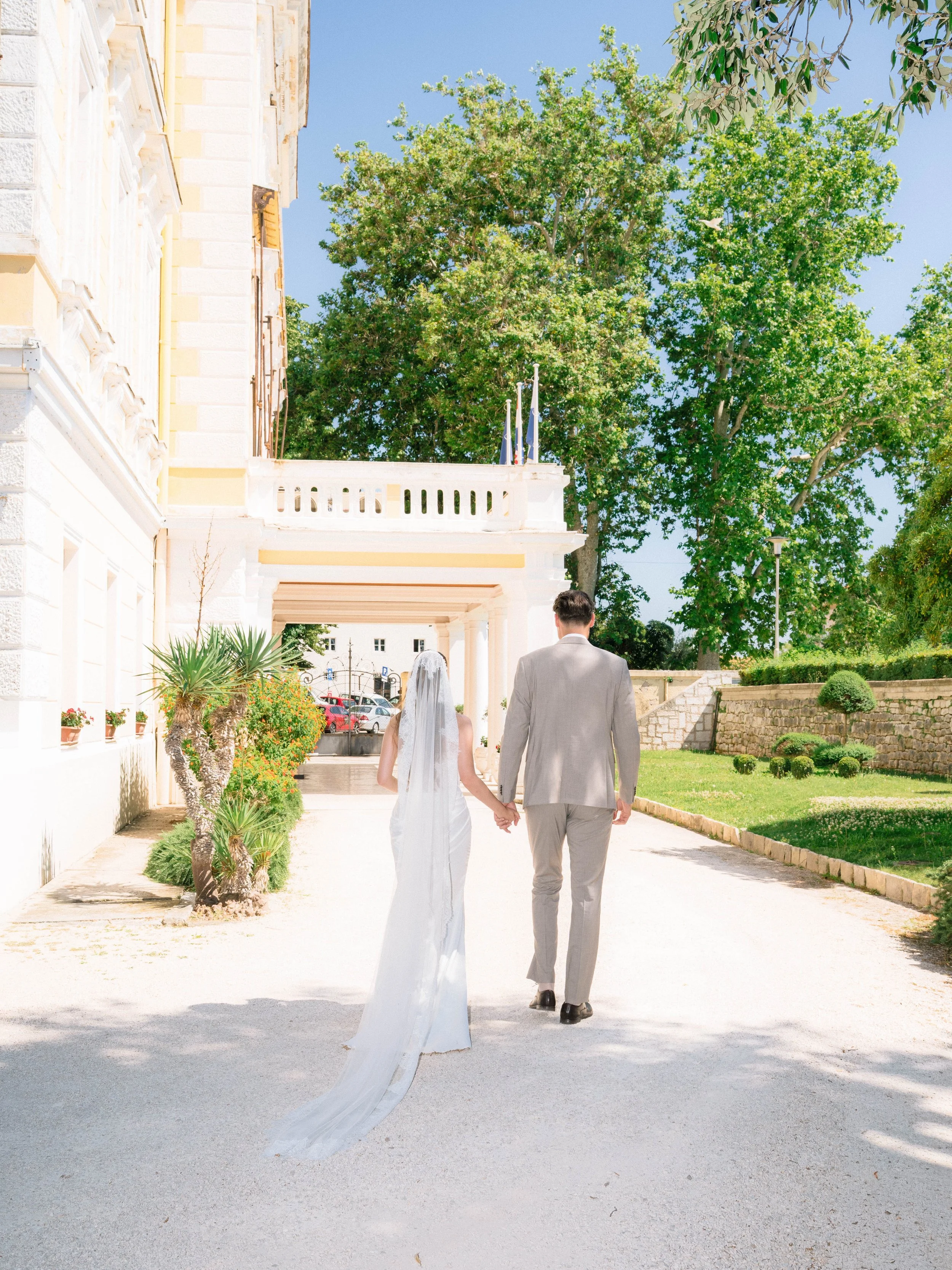 A bride and groom holding hands and walking outdoors on a sunny day, with lush green trees and a light-colored building in the background.