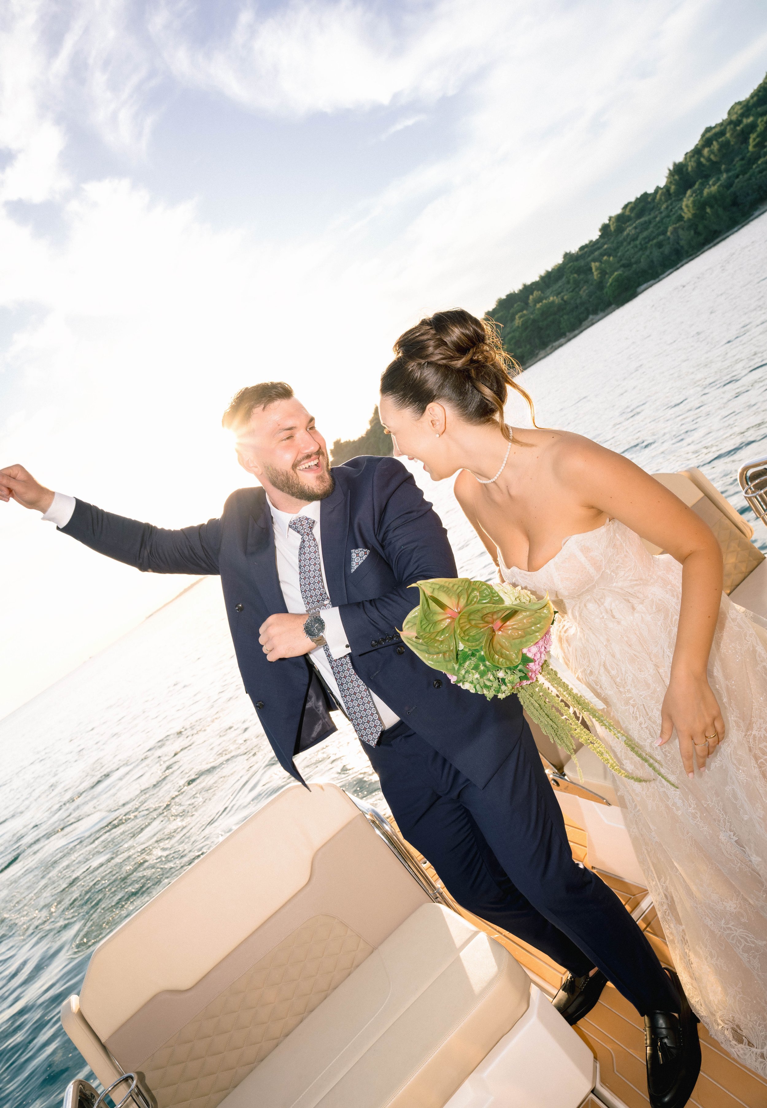 A newlywed couple on a boat, smiling and enjoying a moment together near a body of water with green trees in the background. The groom is dressed in a navy suit with a patterned tie, and the bride is in a white wedding dress holding a bouquet.