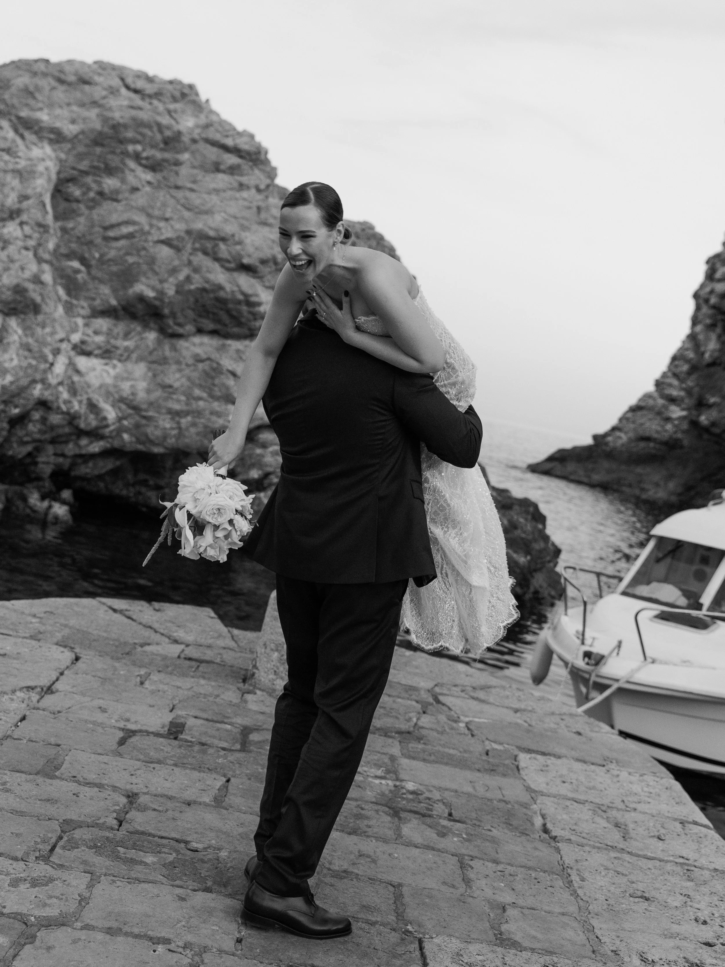A couple wearing wedding attire, with the bride on the groom's back, standing on a stone pathway near a body of water with rocks and a boat in the background, smiling and laughing.
