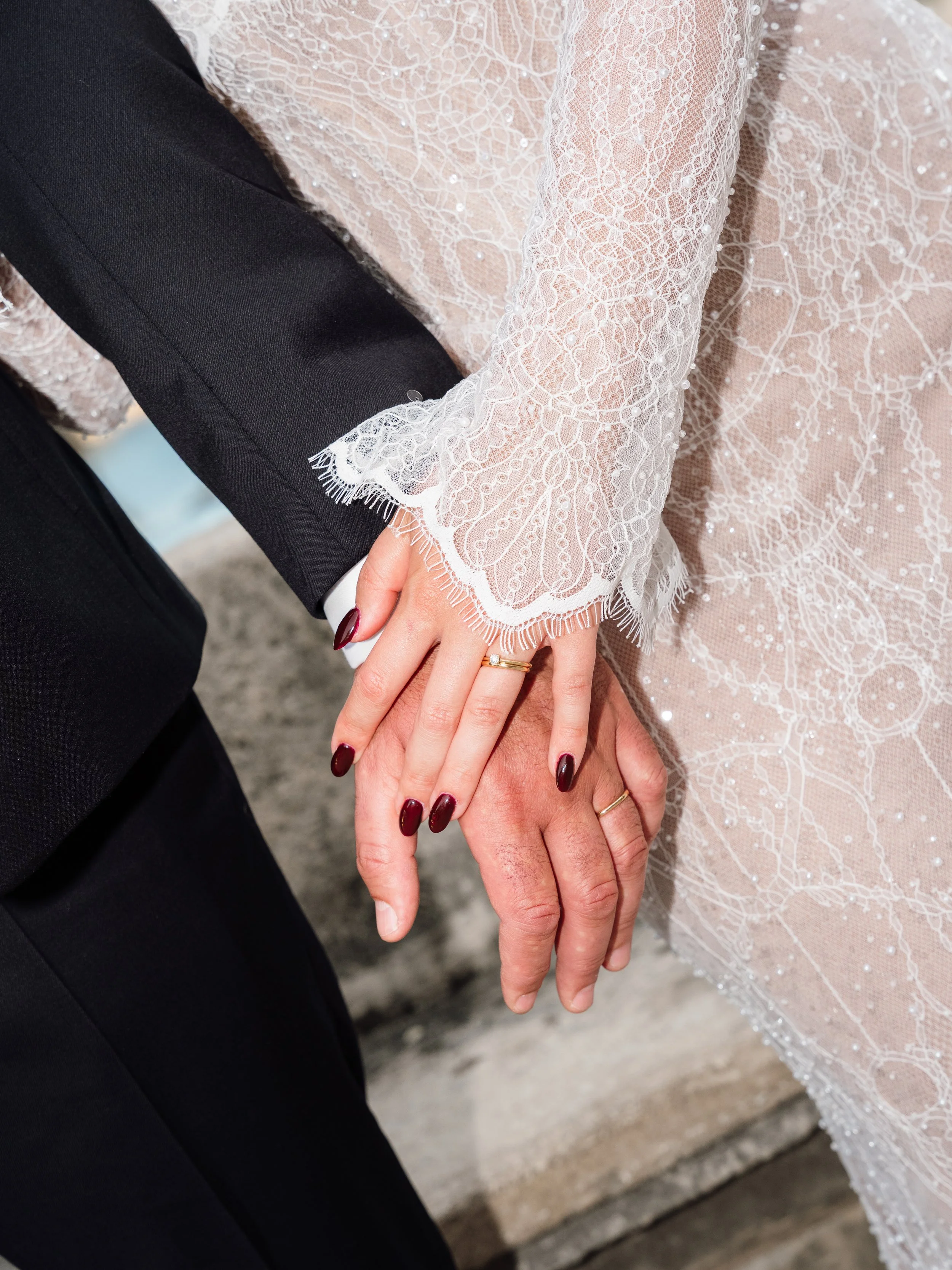 Close-up of a couple holding hands, with the woman wearing a lace wedding dress with intricate details and pearls, and the man wearing a black suit.