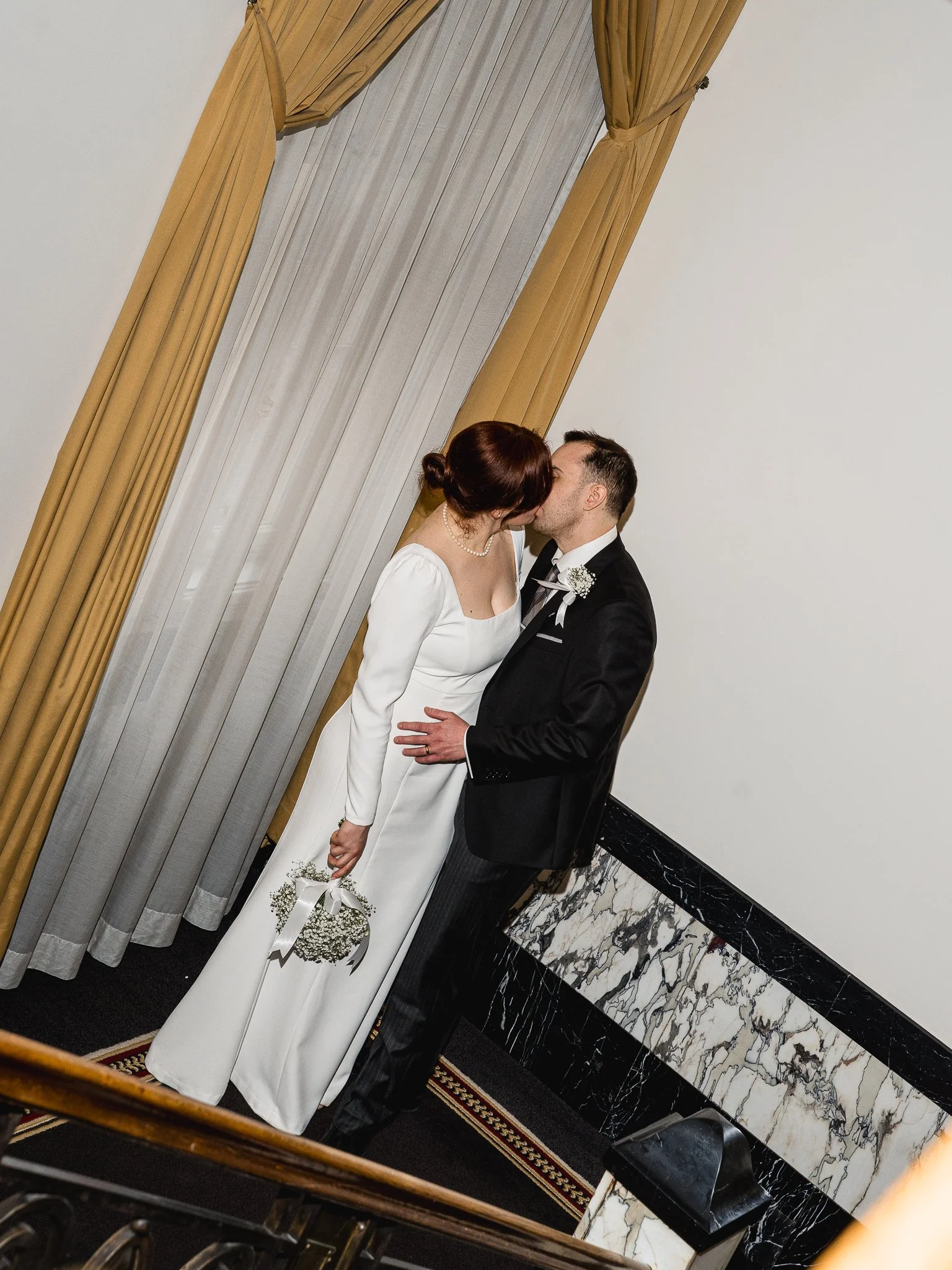 A bride and groom sharing a kiss indoors, with the bride holding a small bouquet of baby's breath flowers. The groom is dressed in a black tuxedo, and the bride is wearing a white wedding dress with long sleeves. They are standing next to a window wi