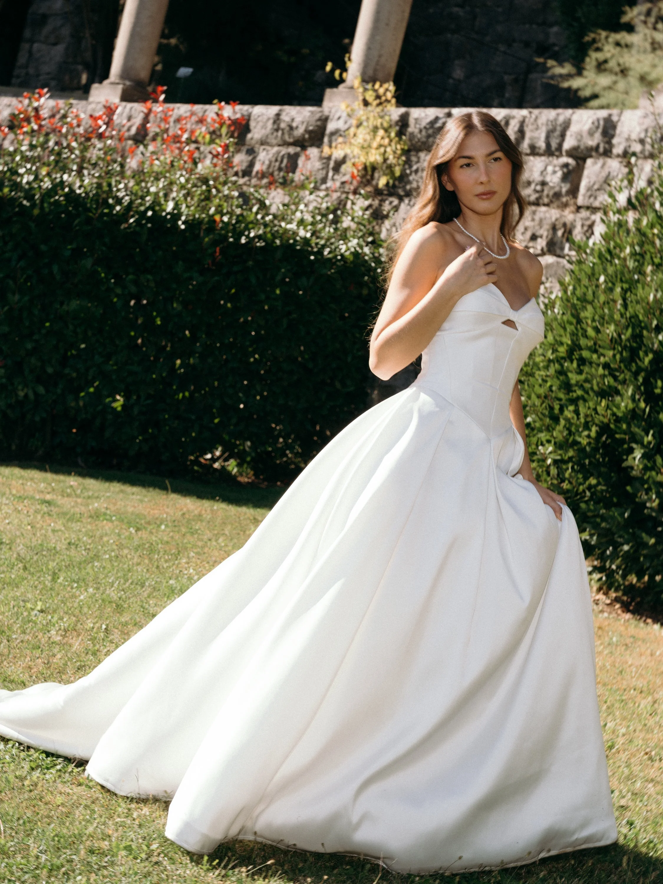 A woman wearing a white wedding dress with a fitted bodice and full skirt, standing outdoors on a grassy area with bushes and stones in the background.
