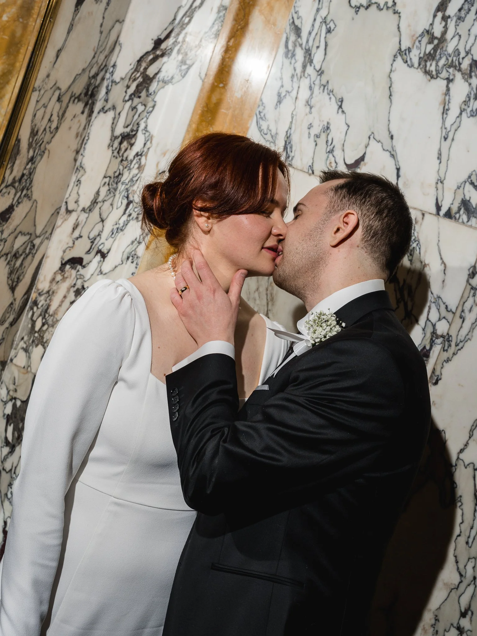 A bride and groom sharing a kiss at a wedding, with the bride wearing a white dress and the groom in a black suit, in front of a marble wall.