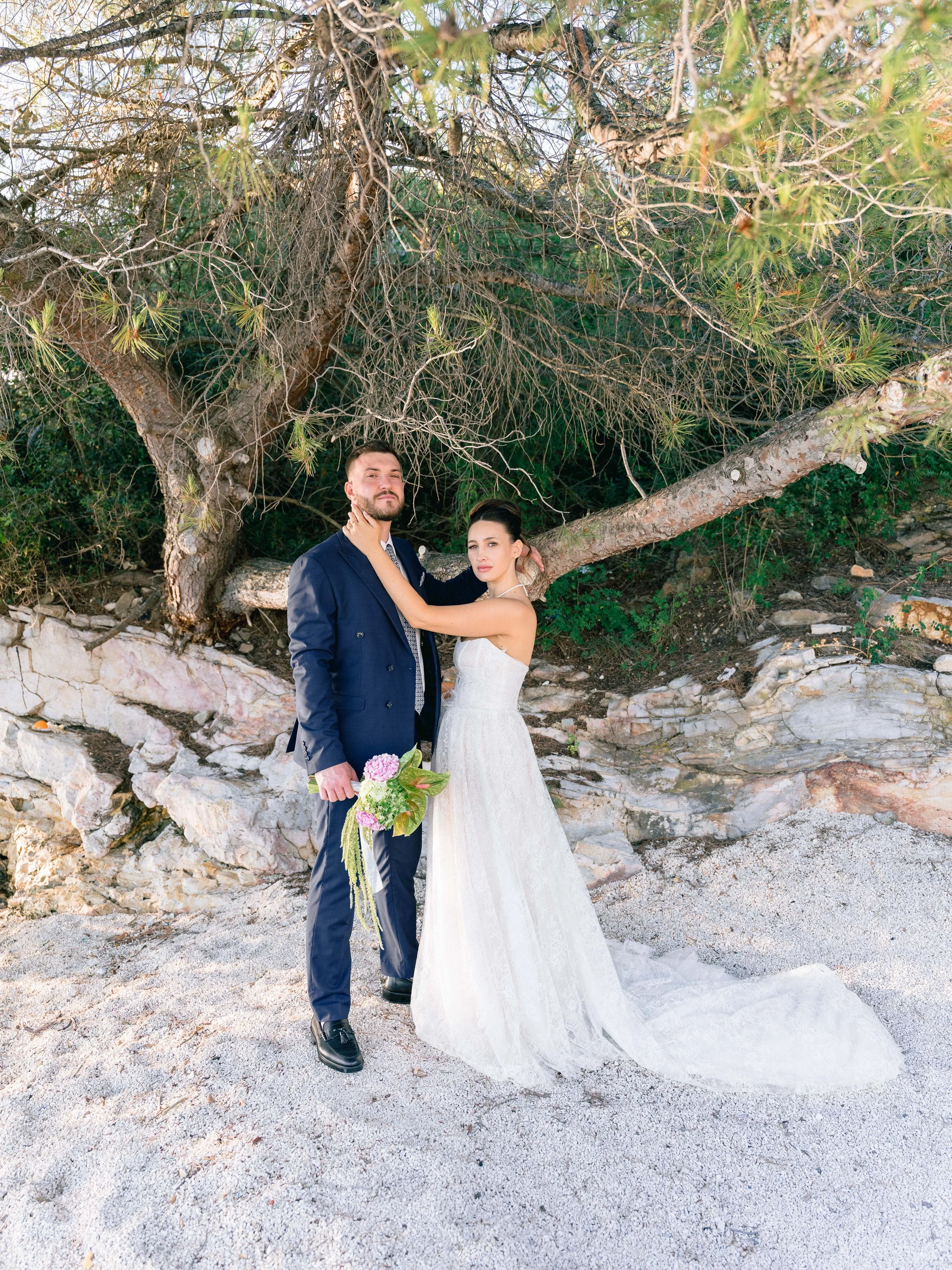 A bride and groom standing on a sandy beach in front of a large tree with sprawling branches and green foliage, posing for a wedding photo.
