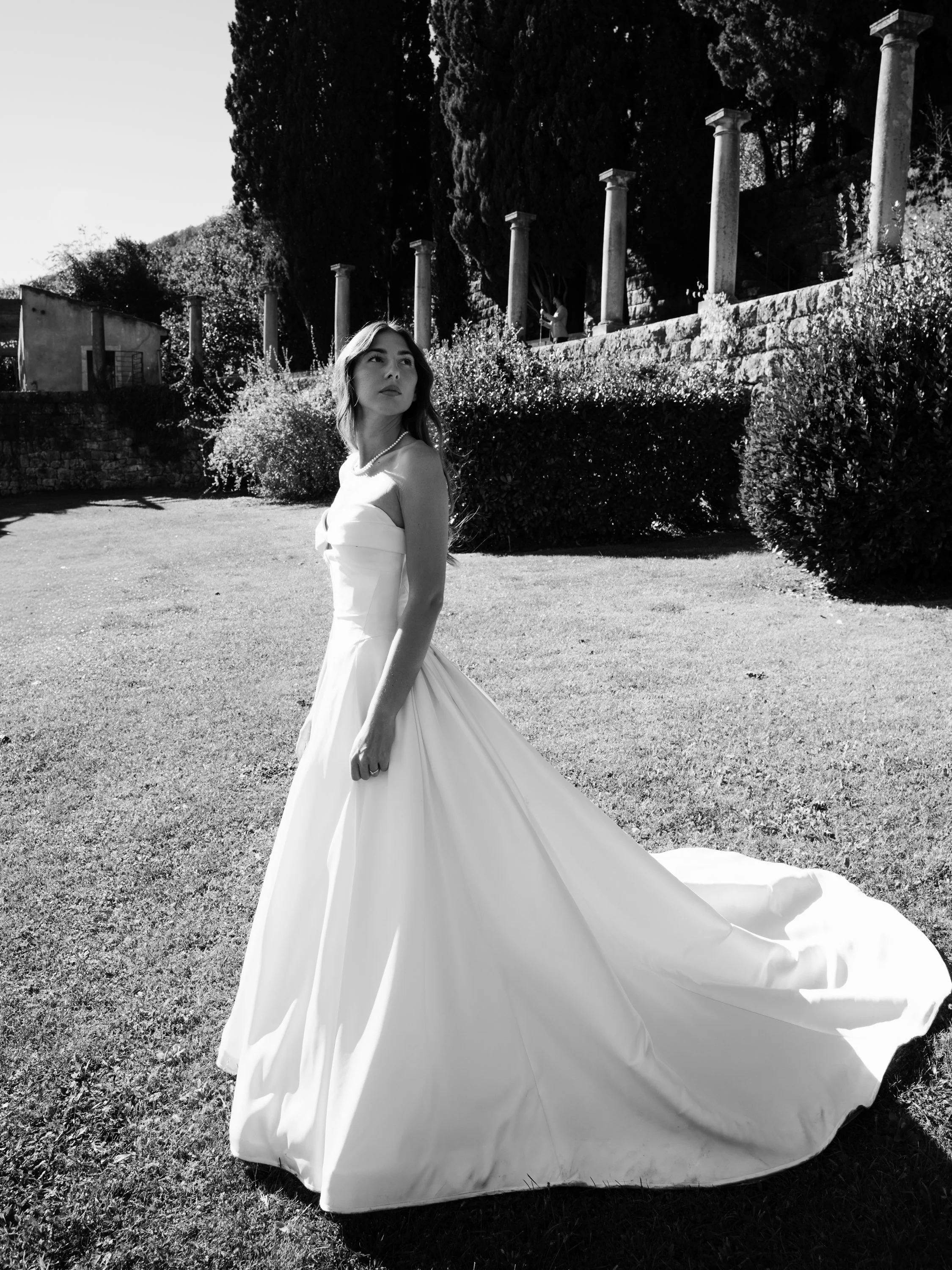 A woman in a white wedding dress standing on a grassy lawn with trees, bushes, and columns in the background, captured in black and white.