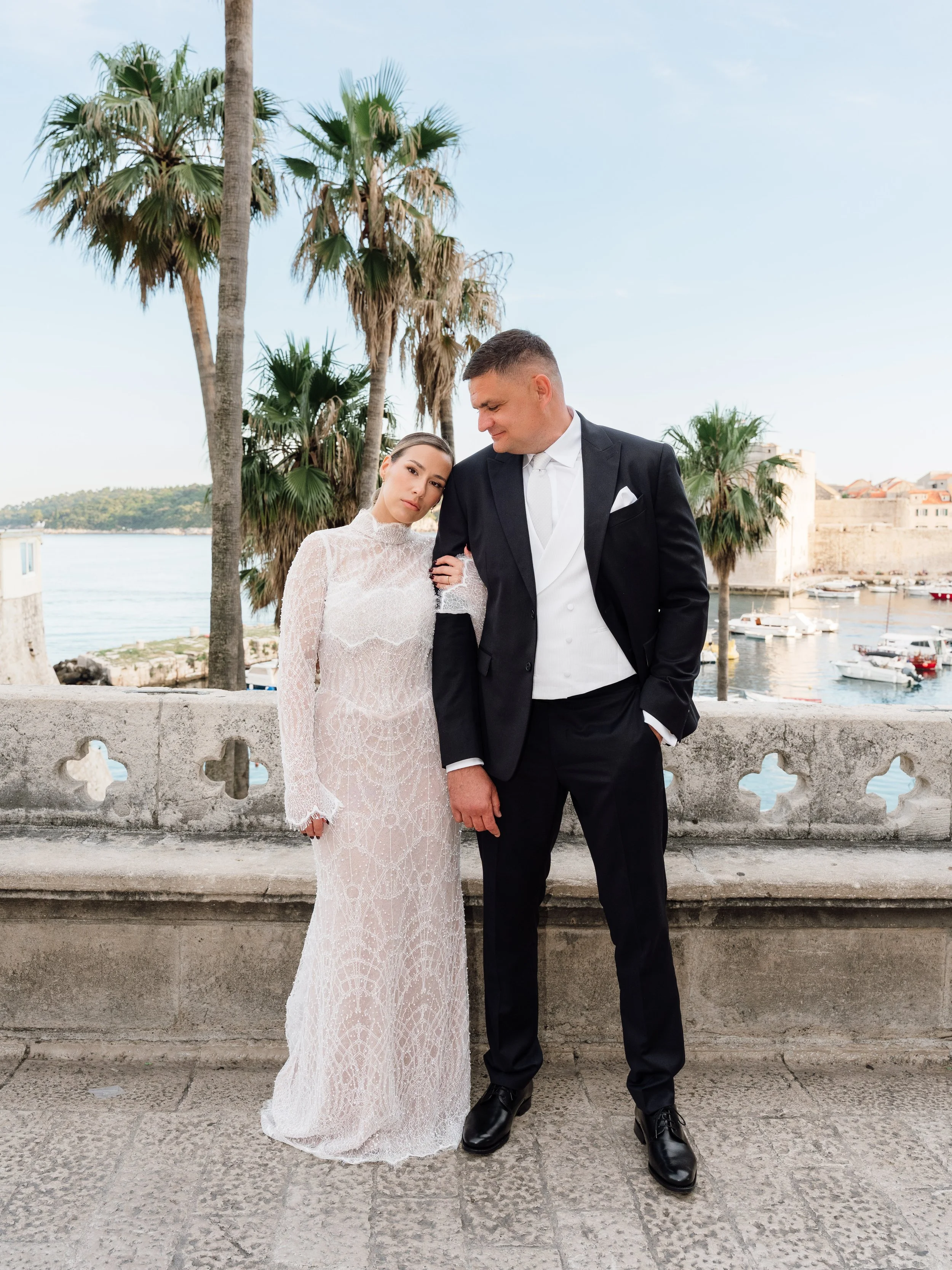 An editorial photo of a bride and groom in formal wedding attire standing by a stone railing with palm trees and a harbor in the background in in Dubrovnik, Croatia.