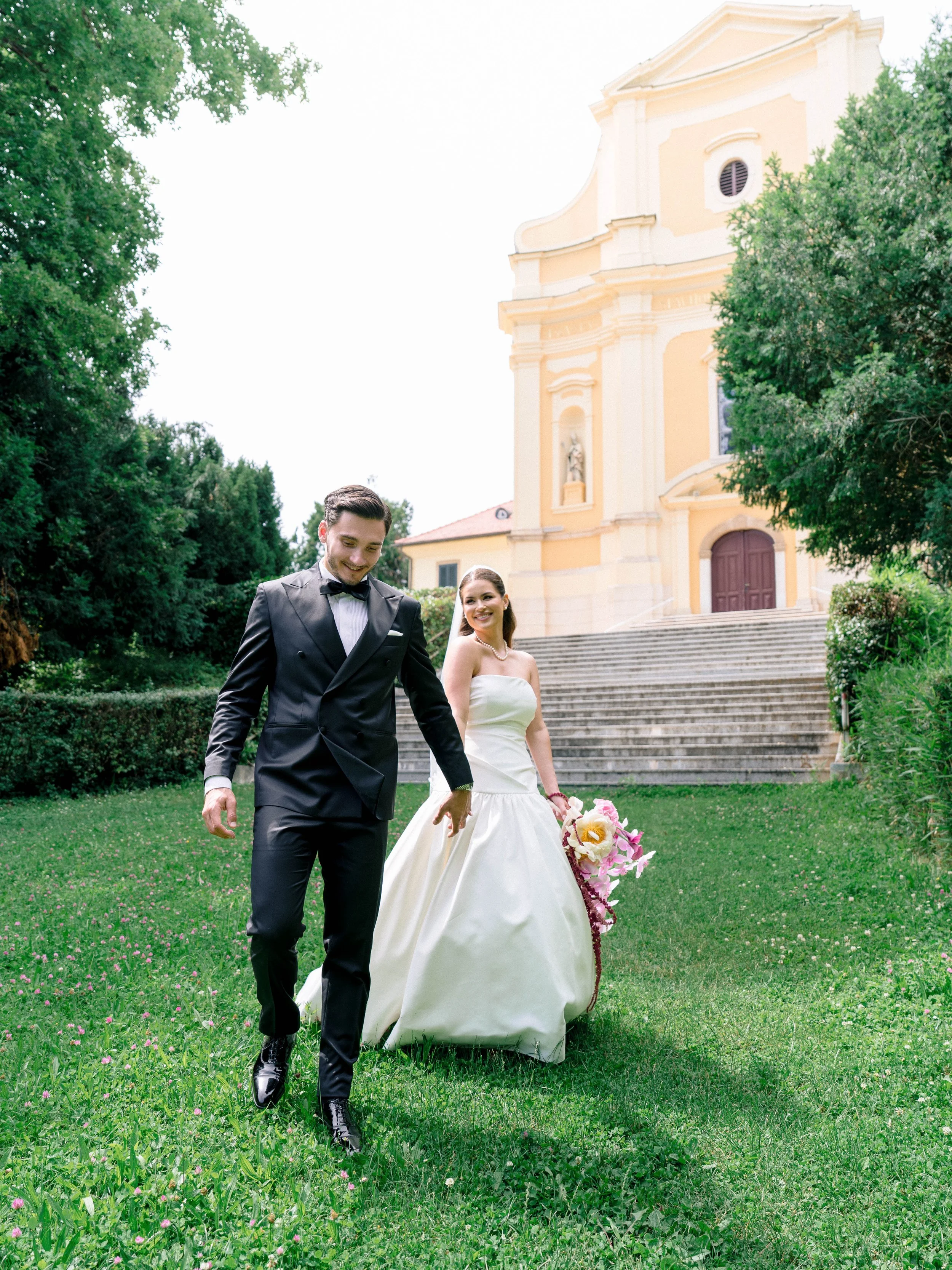 A bride and groom walking in a park in front of a yellow church, with steps leading up to its entrance, surrounded by green trees and grass.