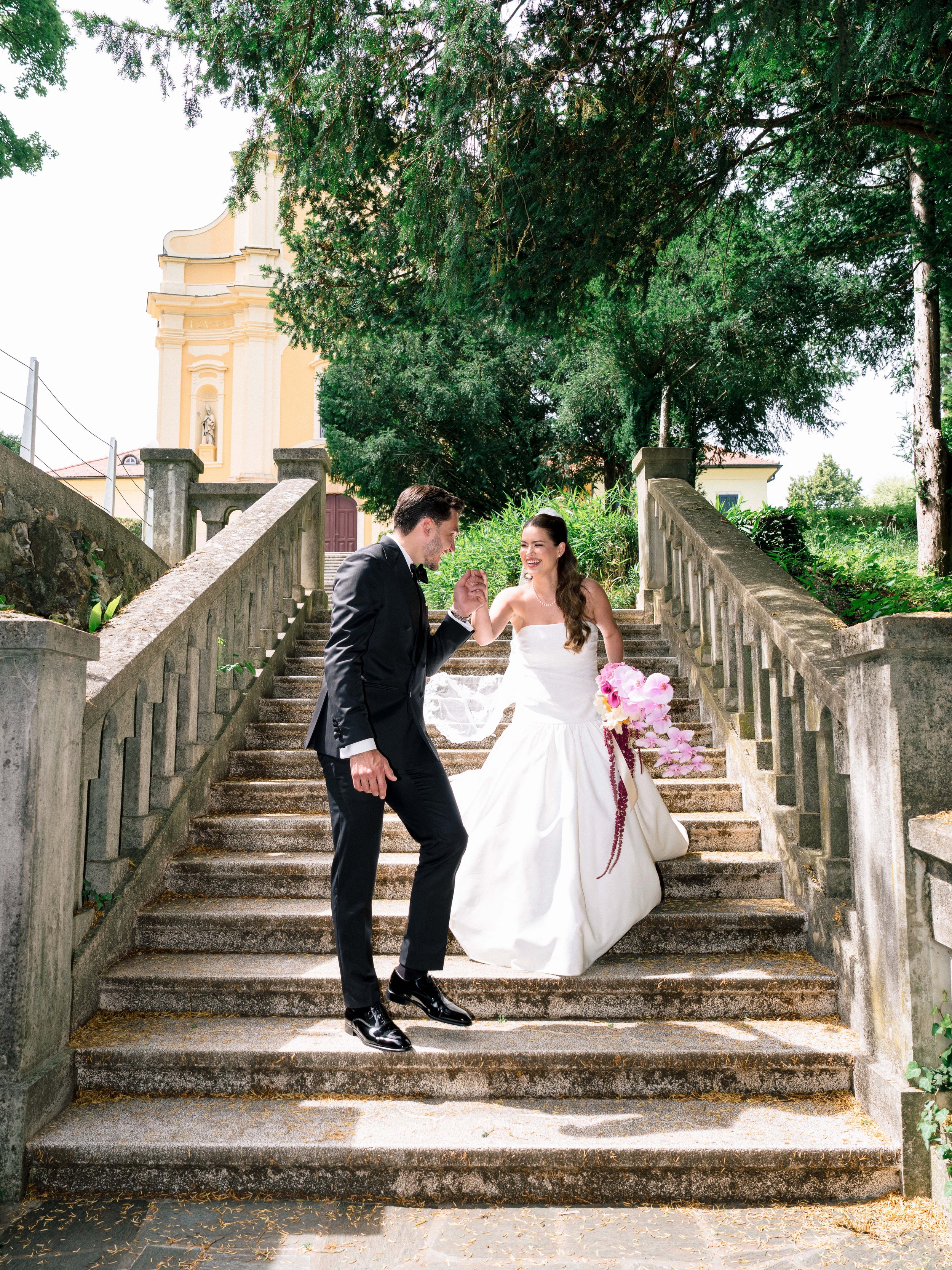 A bride and groom dancing on outdoor stone staircase surrounded by greenery with a church in the background.