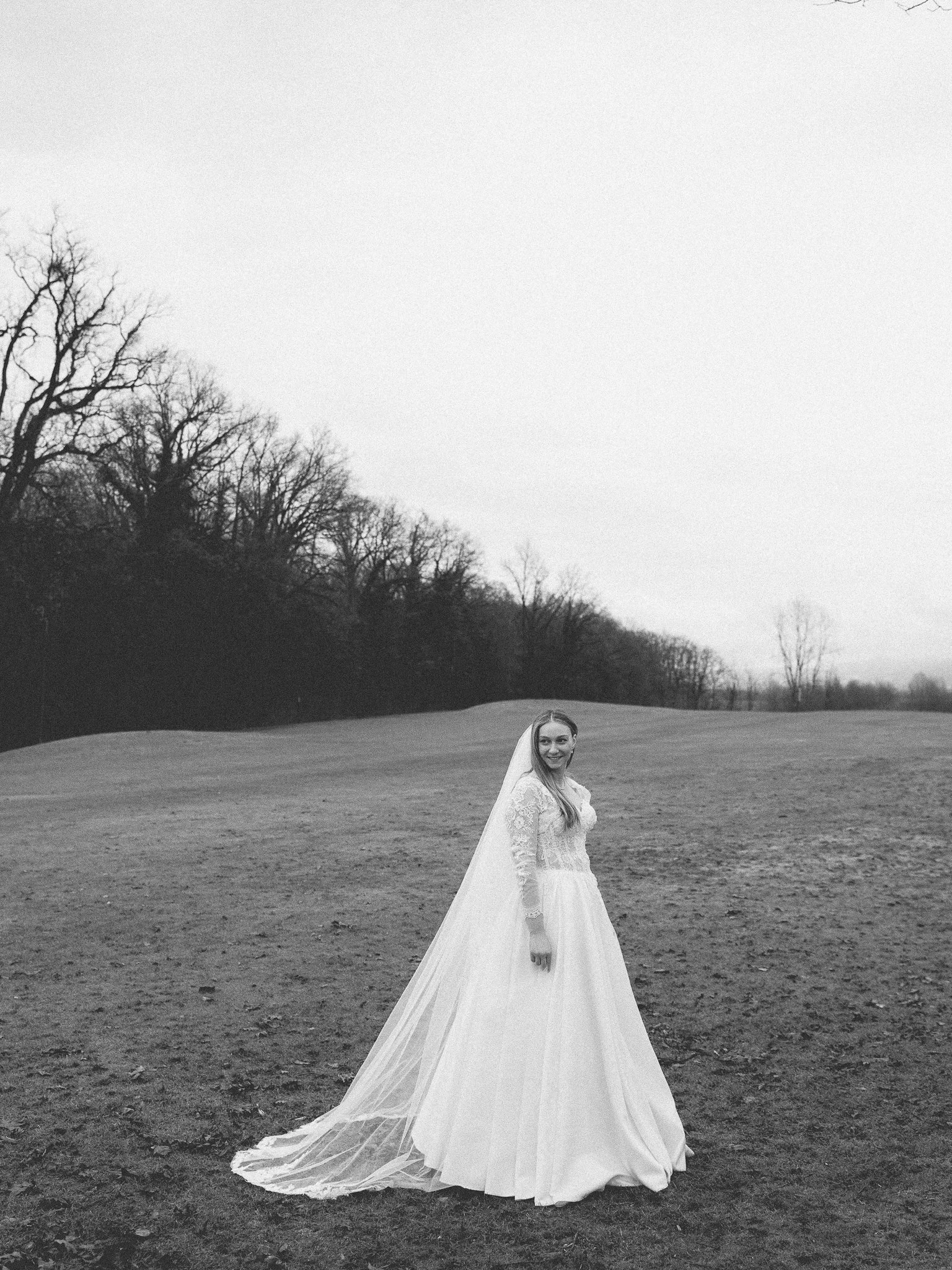 Black and white photo of a bride in a wedding dress with a long train standing in an open field with leafless trees in the background.