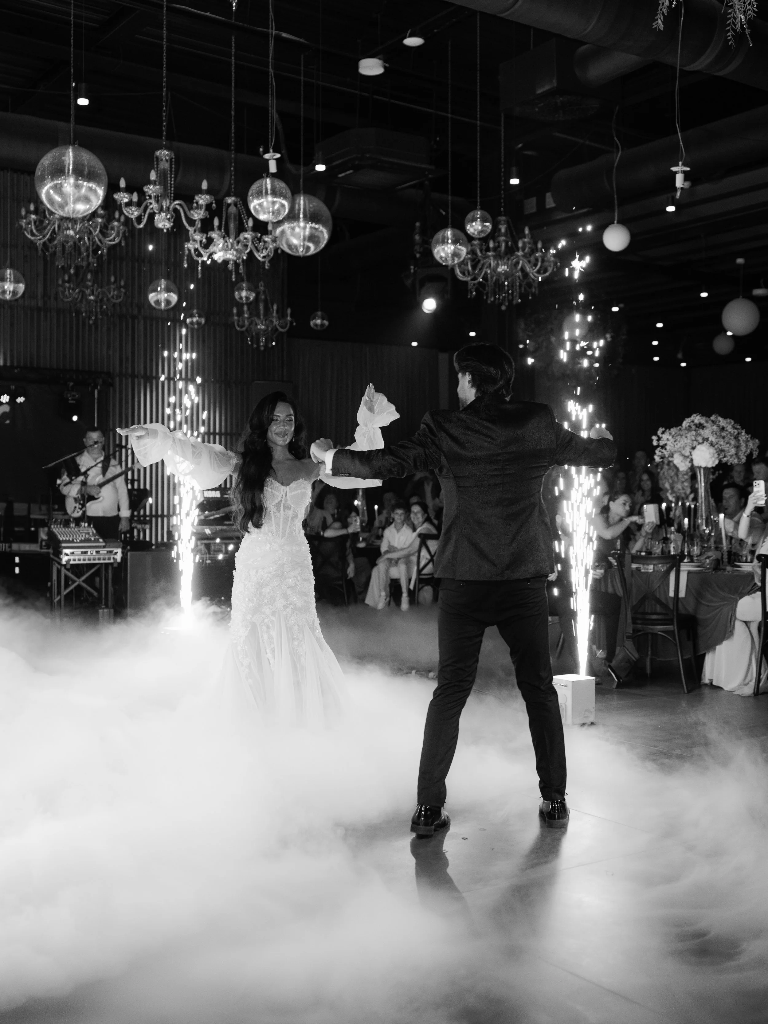 A bride and groom dancing at their wedding reception, surrounded by guests, with a fog machine, sparklers, and chandeliers hanging from the ceiling.