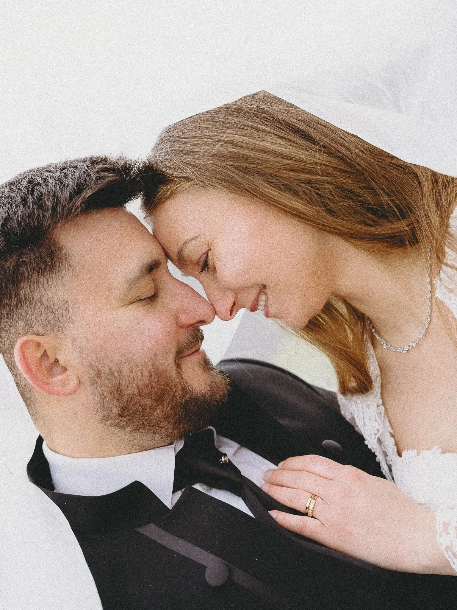 A close-up of a couple on their wedding day, with their foreheads and noses touching and eyes closed, smiling gently. The woman has long, wavy red hair and is wearing a pearl necklace and a wedding dress with lace details. The man has short dark hair