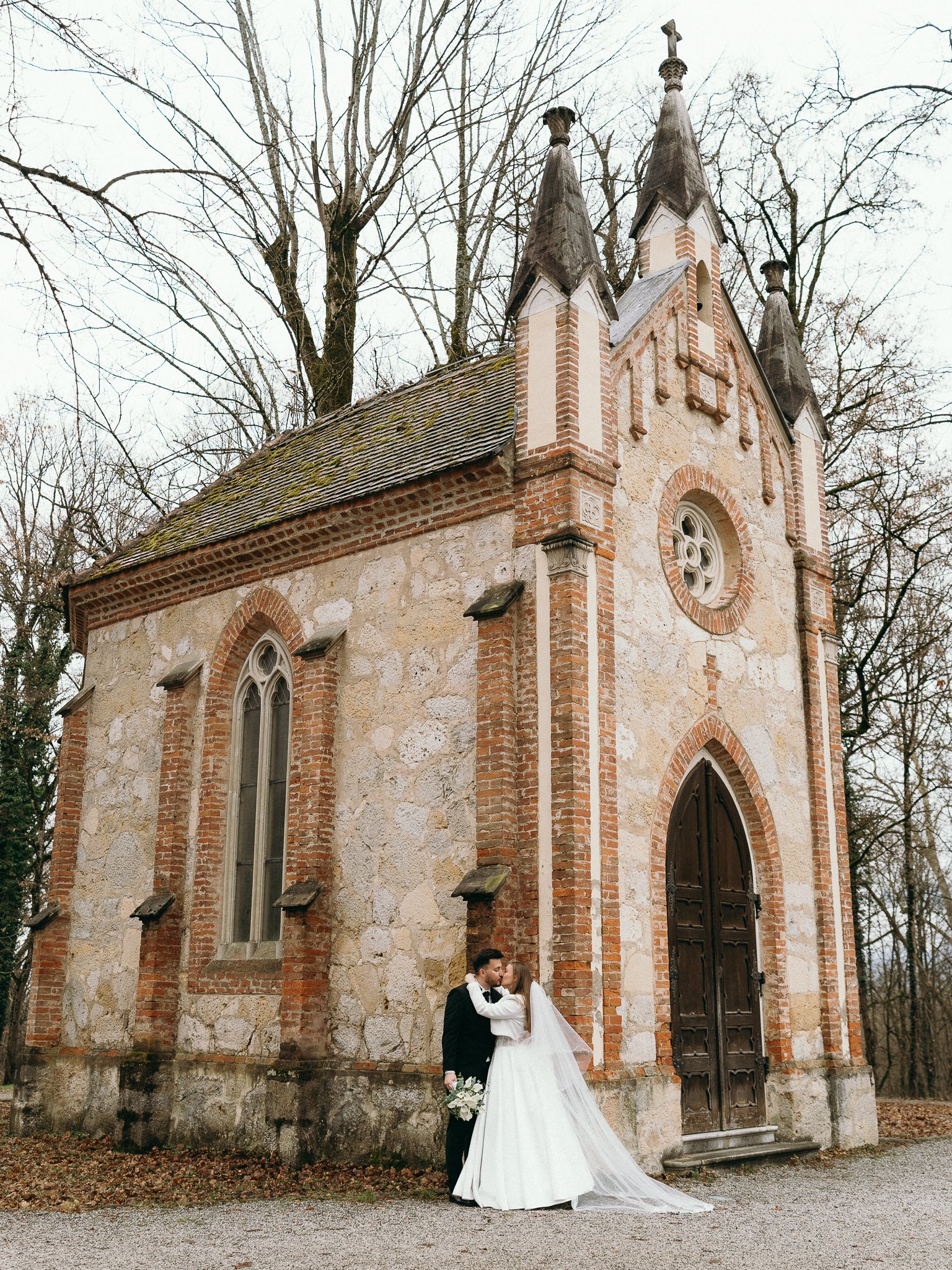 A bride and groom kissing outside a small historic stone and brick chapel, with leafless trees in the background.