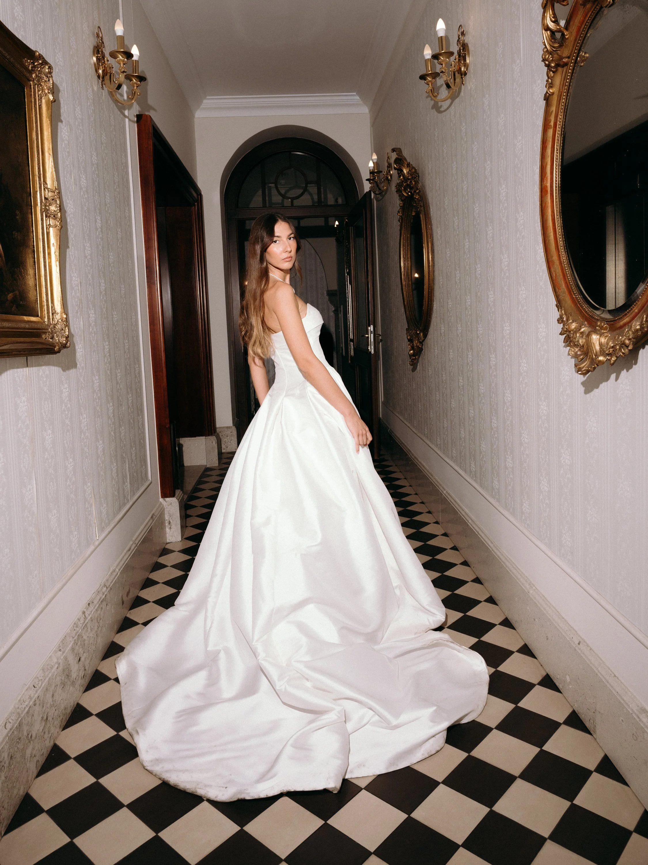 A woman in a white wedding dress standing in a hallway with checkered black and white flooring, ornate gold-framed mirrors, and vintage wall sconces.