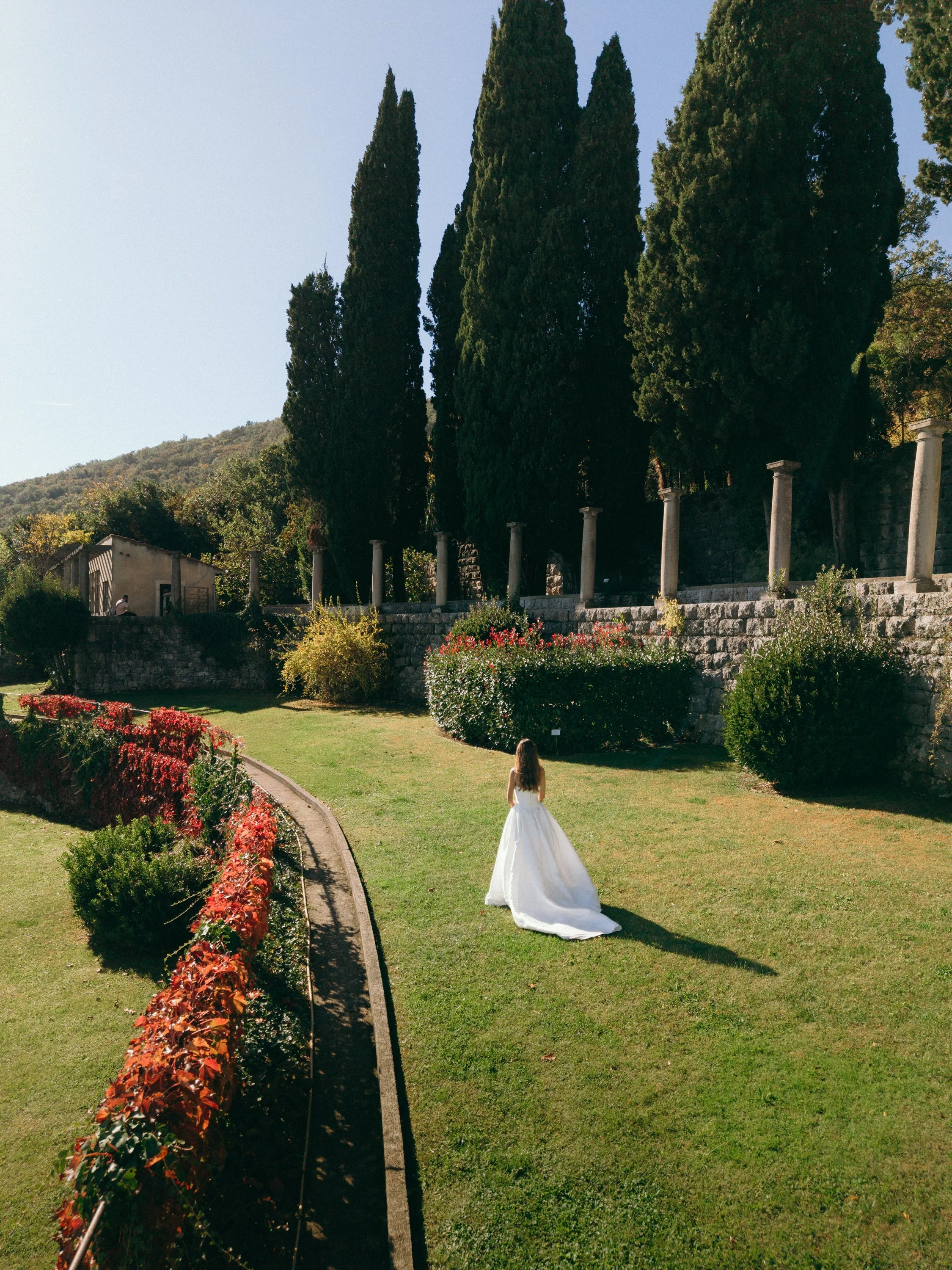 A woman in a white wedding dress walking on a well-maintained grass lawn, bordered by colorful bushes and trees, with tall cypress trees and a stone wall with columns in the background.