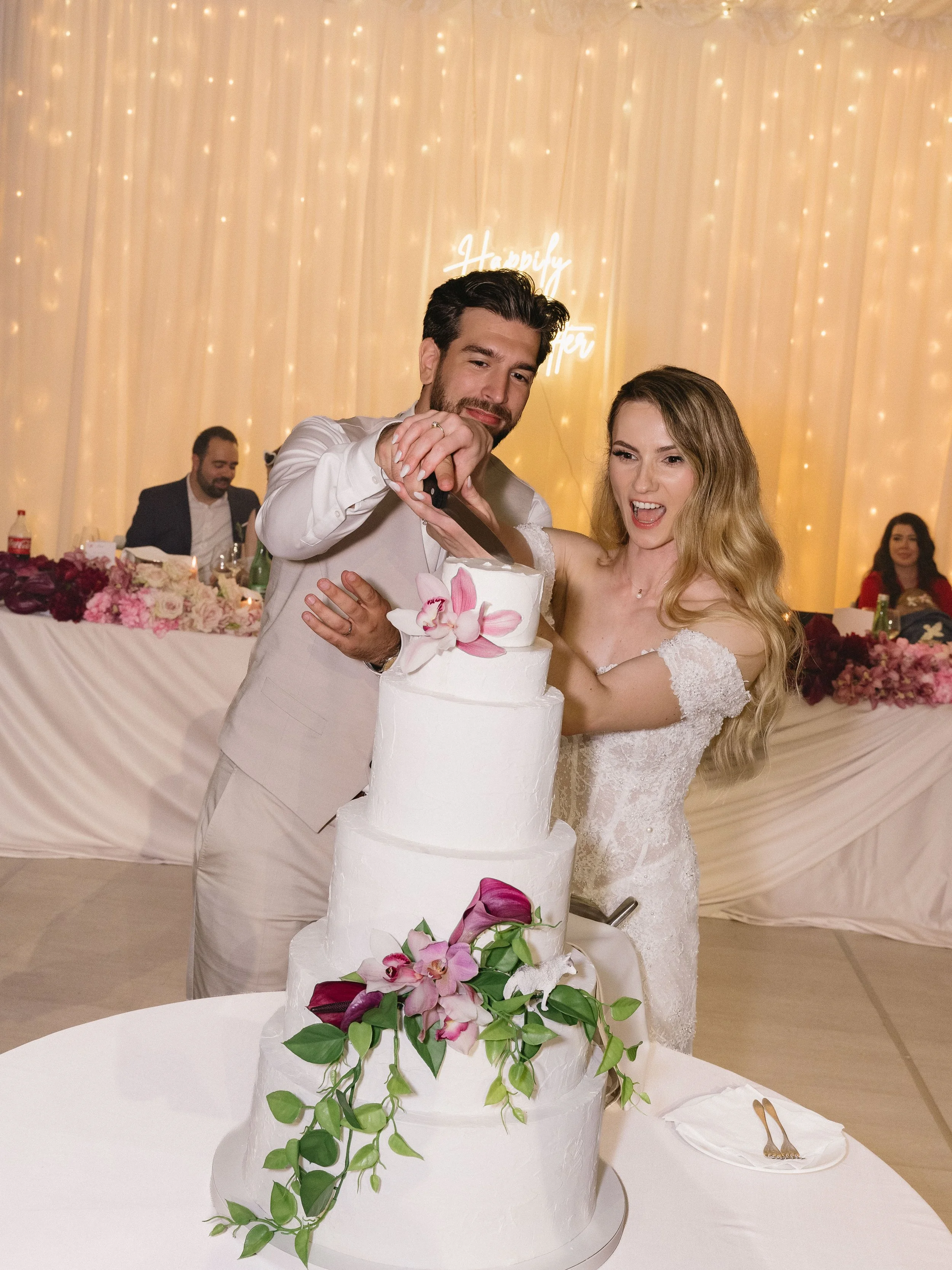 A bride and groom cut a three-tier white wedding cake decorated with pink and purple orchids and green leaves, during a wedding celebration in a decorated hall.
