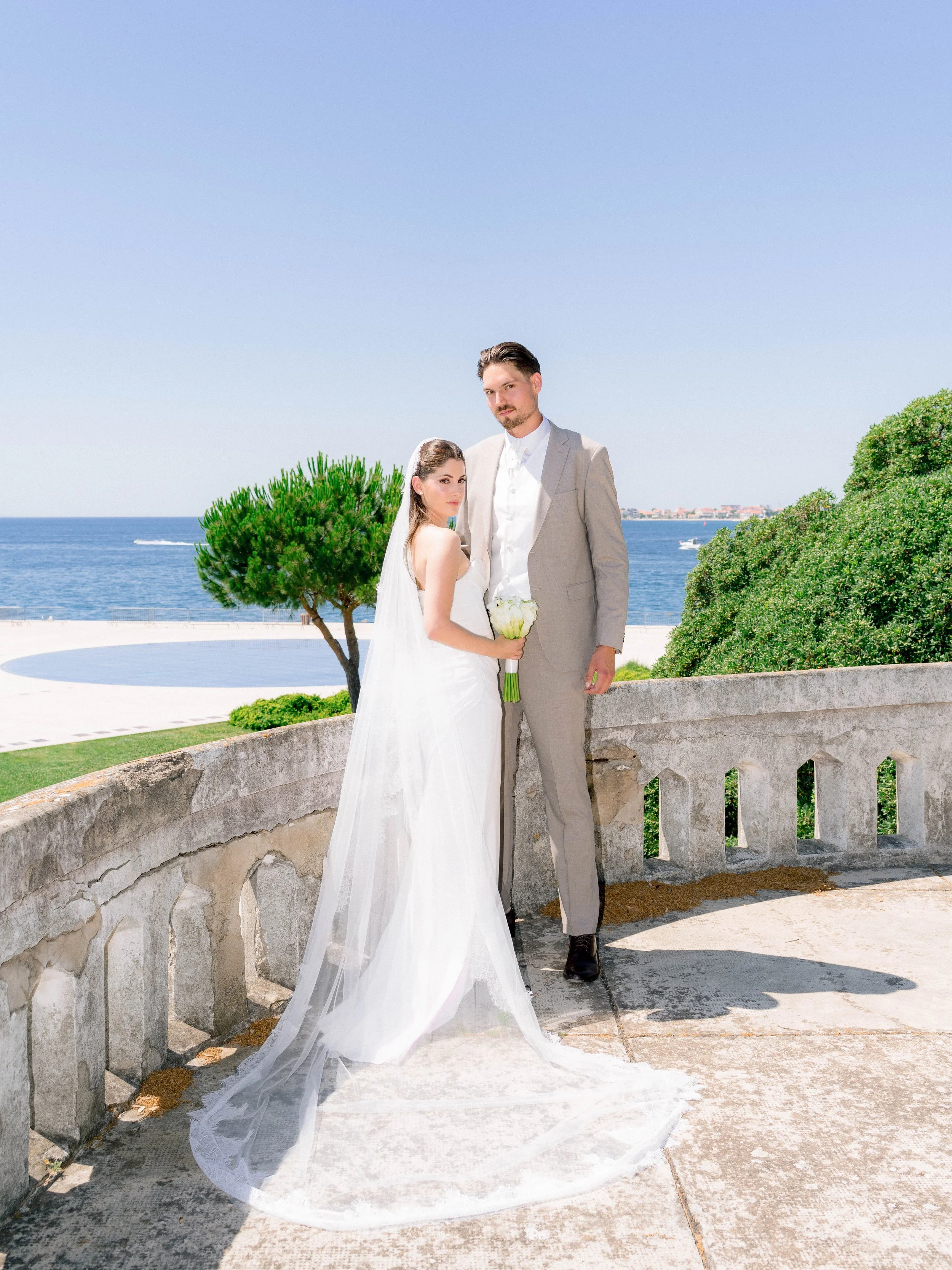 A bride and groom standing on a stone balcony near the ocean, with a tree and green bushes in the background, during daytime.