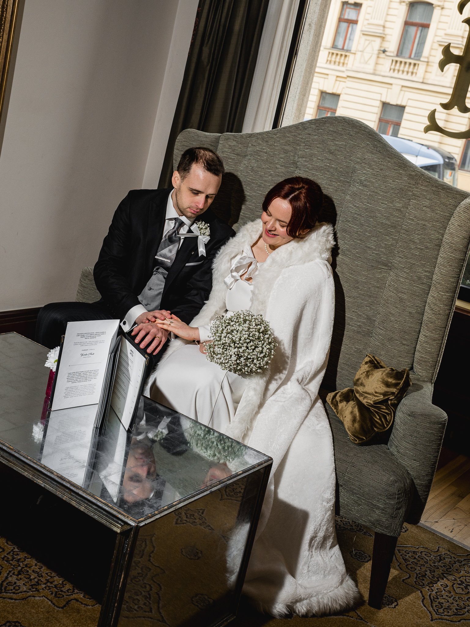 A bride and groom sitting on a high-backed couch inside a building, holding hands, with the bride smiling and holding a bouquet of flowers, and a window behind them showing a city street.