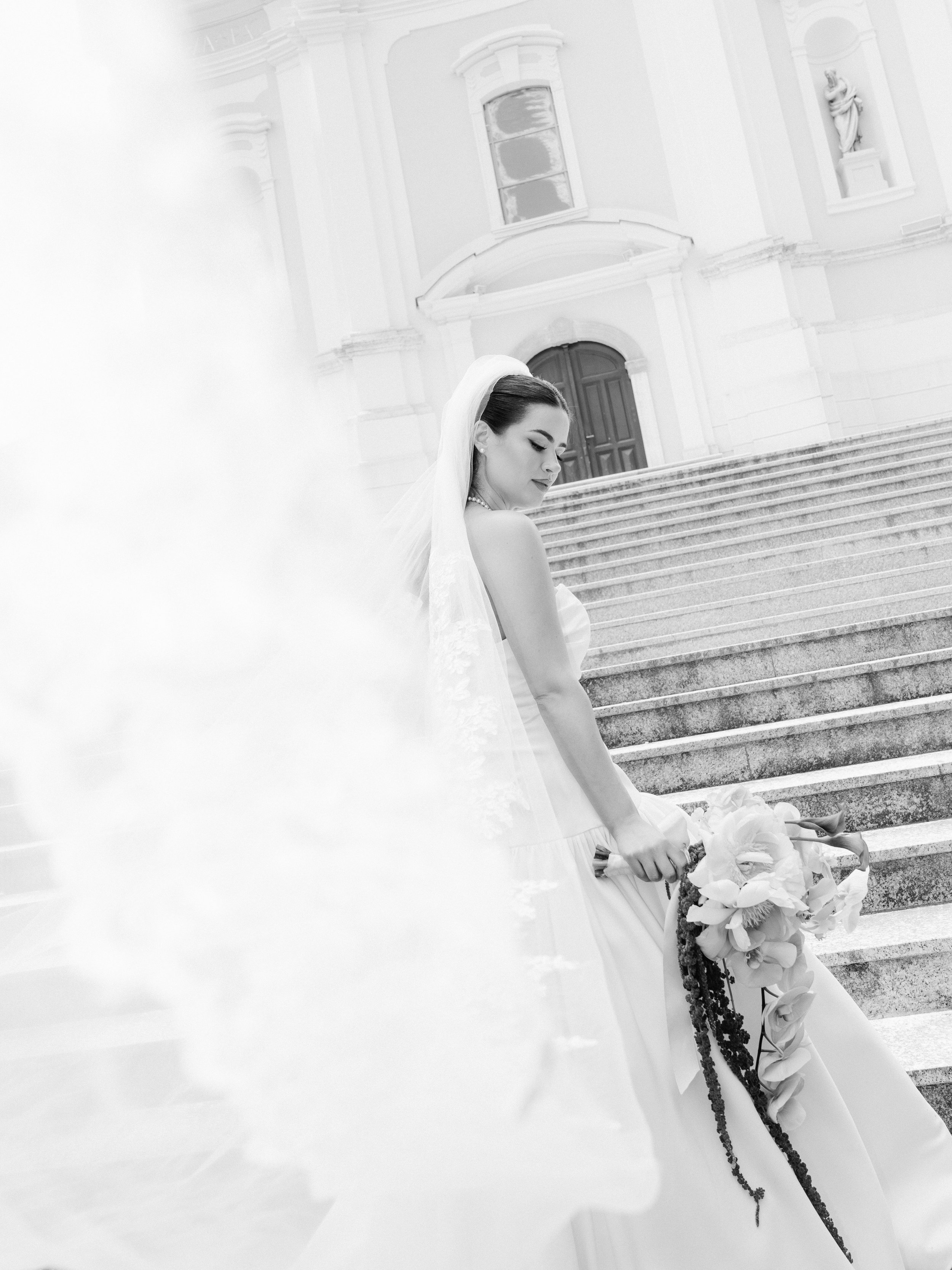 Black and white photo of a bride in a wedding dress holding a bouquet, standing on stone steps in front of a historic building with large arched door and statues in niches.