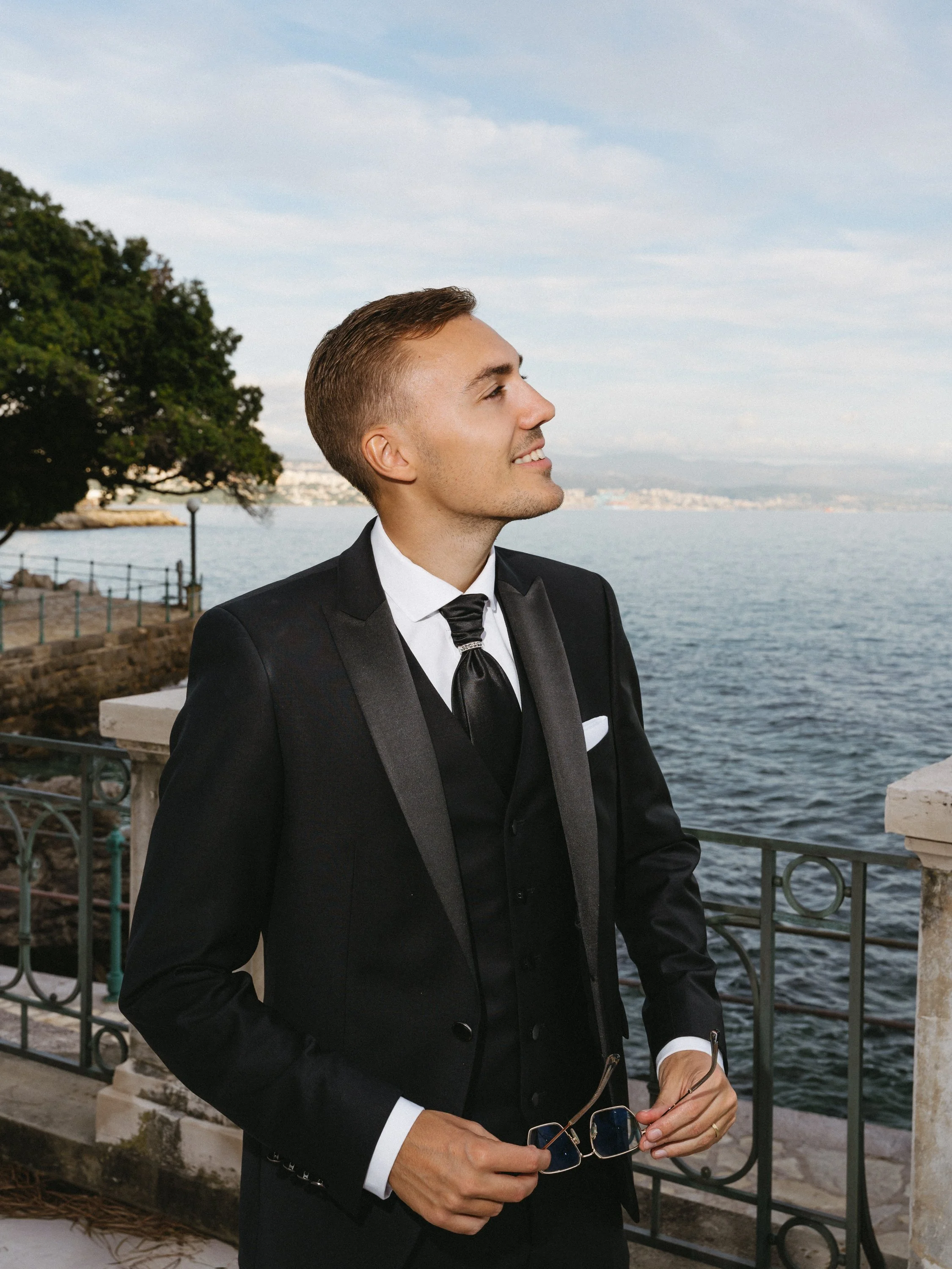 A young man in a black tuxedo standing outdoors by a waterfront, holding sunglasses and enjoying the view.