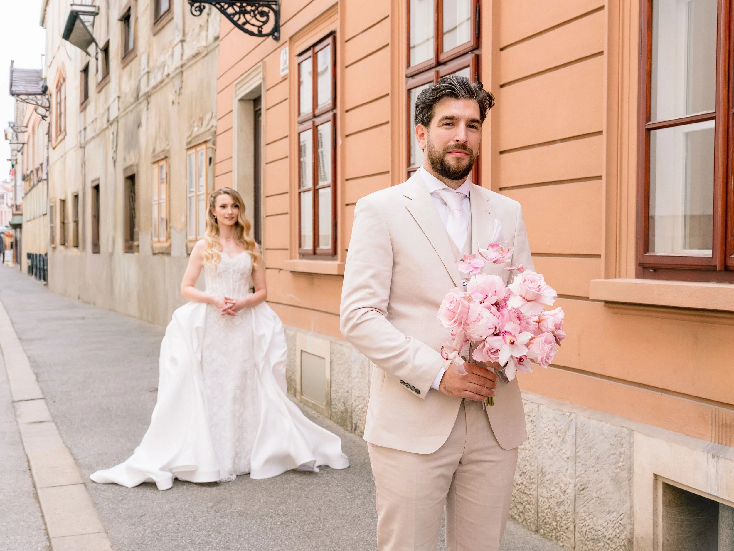 A groom in a white suit holding a bouquet of pink flowers, standing in front of a building with colorful window frames. A bride in a white wedding dress is following behind him on a street.