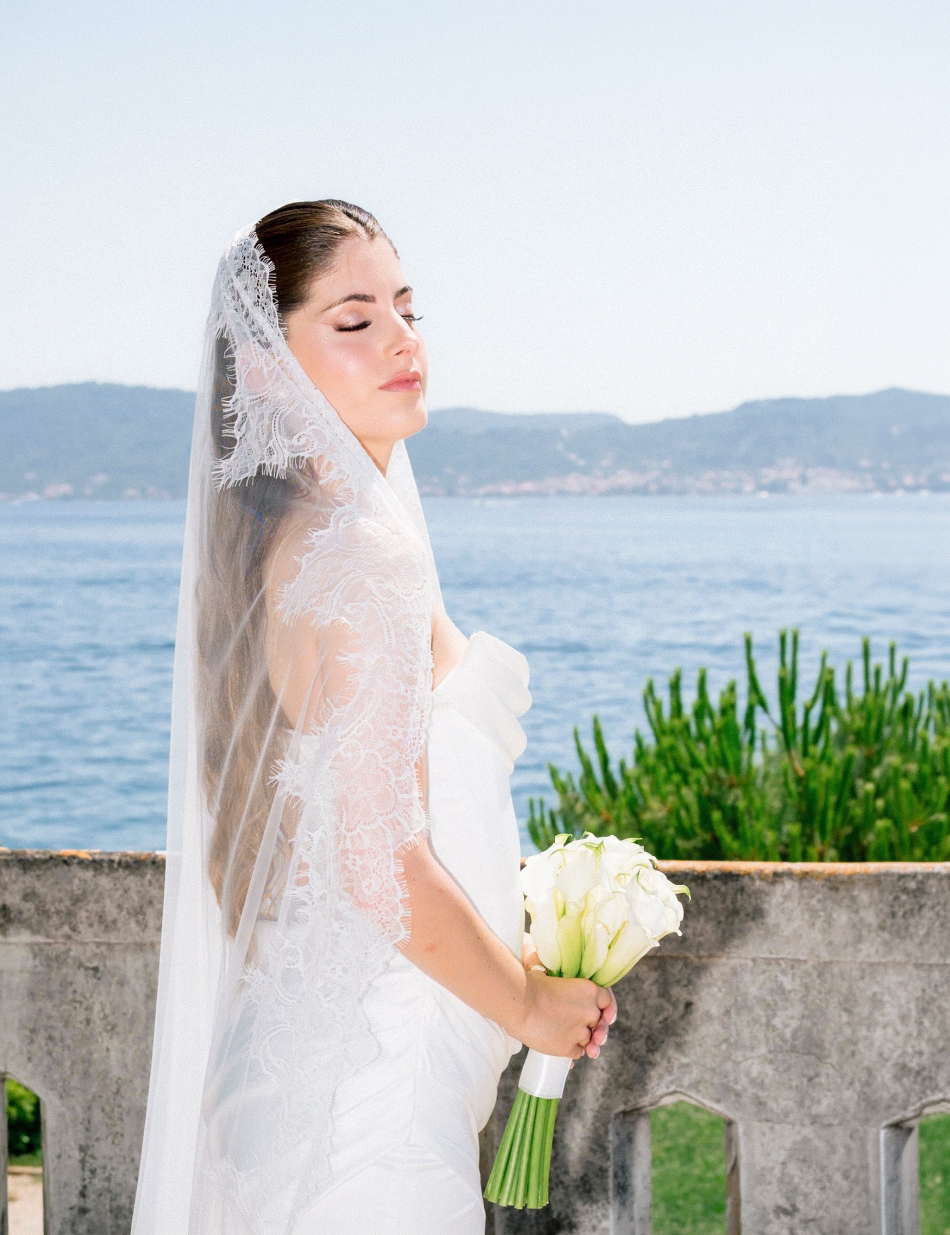 A bride standing outdoors near a stone railing, holding a bouquet of white calla lilies, with her eyes closed and wearing a lace wedding dress with a veil. In the background, there is water and mountains.