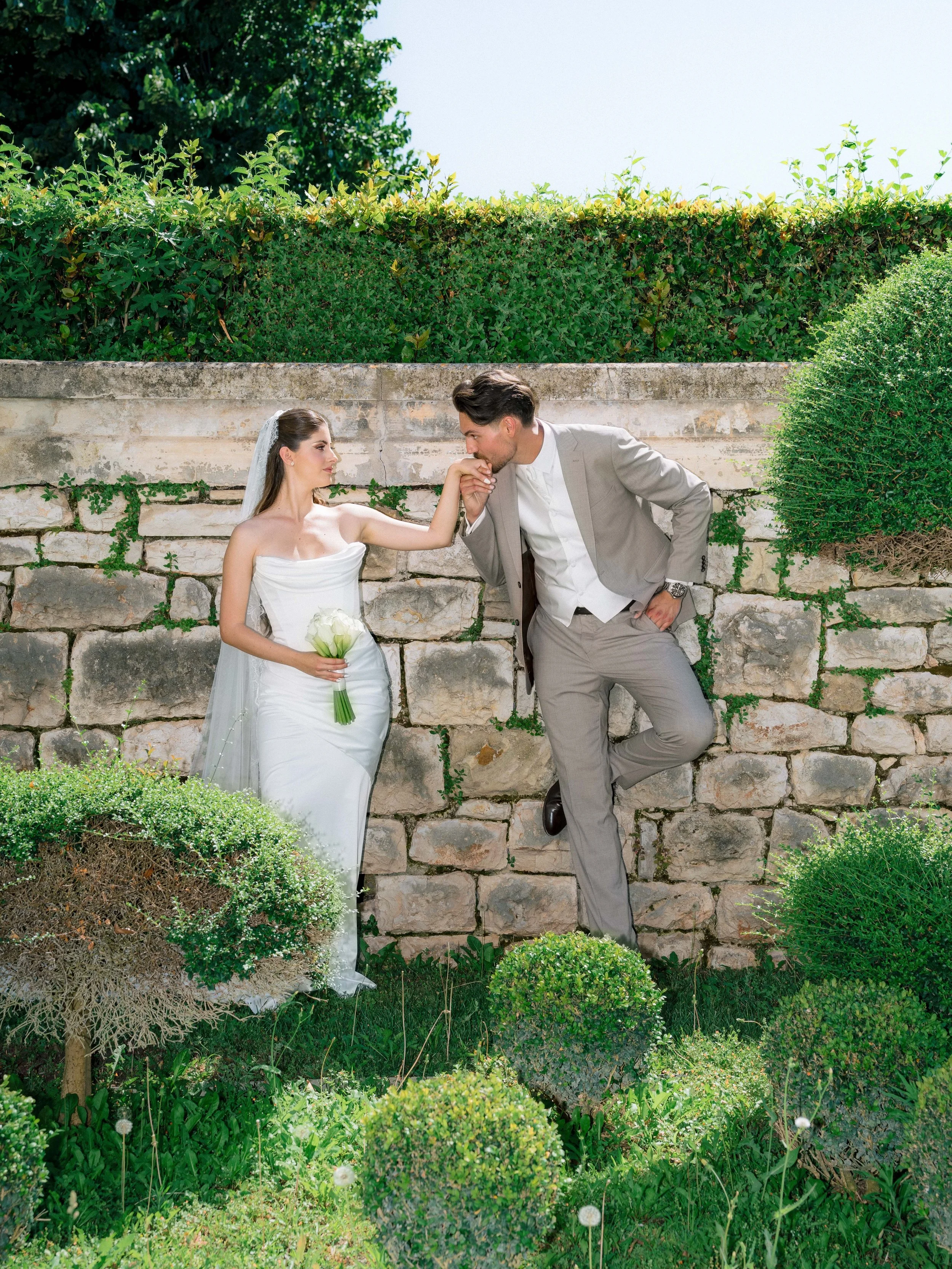 A bride and groom on their wedding day, standing outdoors in front of a stone wall with greenery, the groom gently kissing the bride's hand while she holds a bouquet of white flowers.