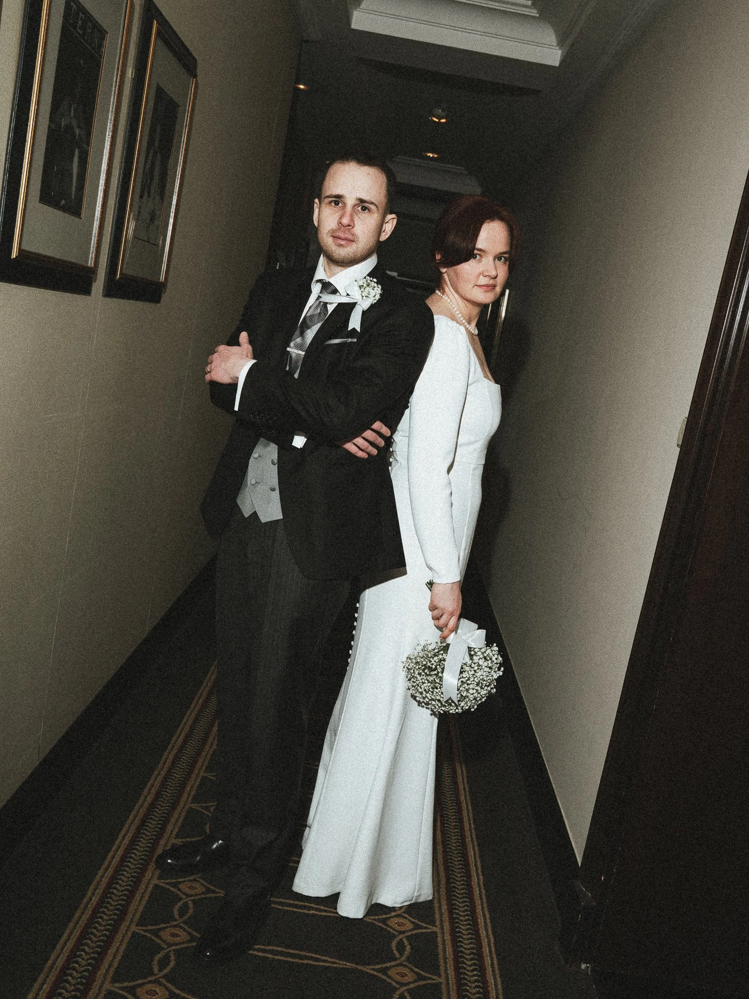 A bride and groom standing back-to-back in a hotel hallway, dressed in wedding attire. The groom wears a black tuxedo with a white shirt and tie, while the bride wears a white wedding gown holding a bouquet of baby's breath.