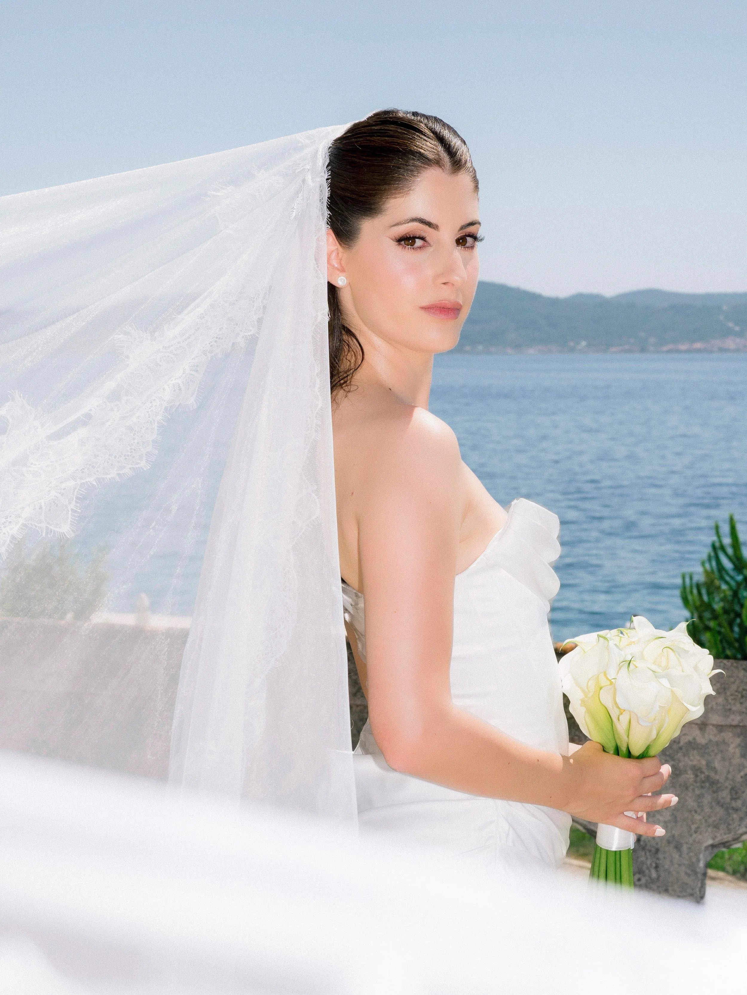 A bride standing outdoors near water, holding a bouquet of white calla lilies, with a sheer lace veil and distant mountains in the background.