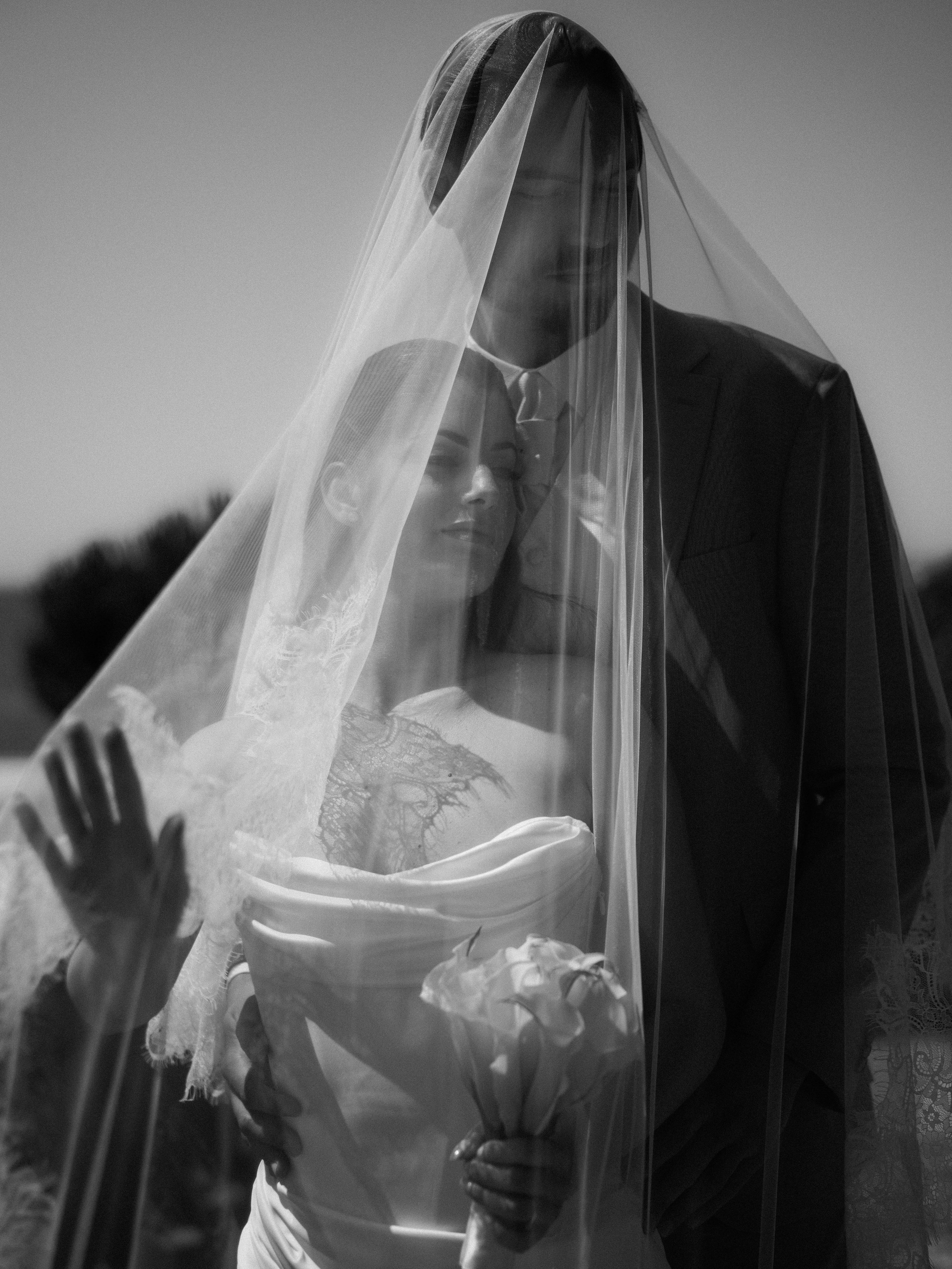 A man and a woman dressed in wedding attire standing behind a window with a veil partially covering the woman's face.