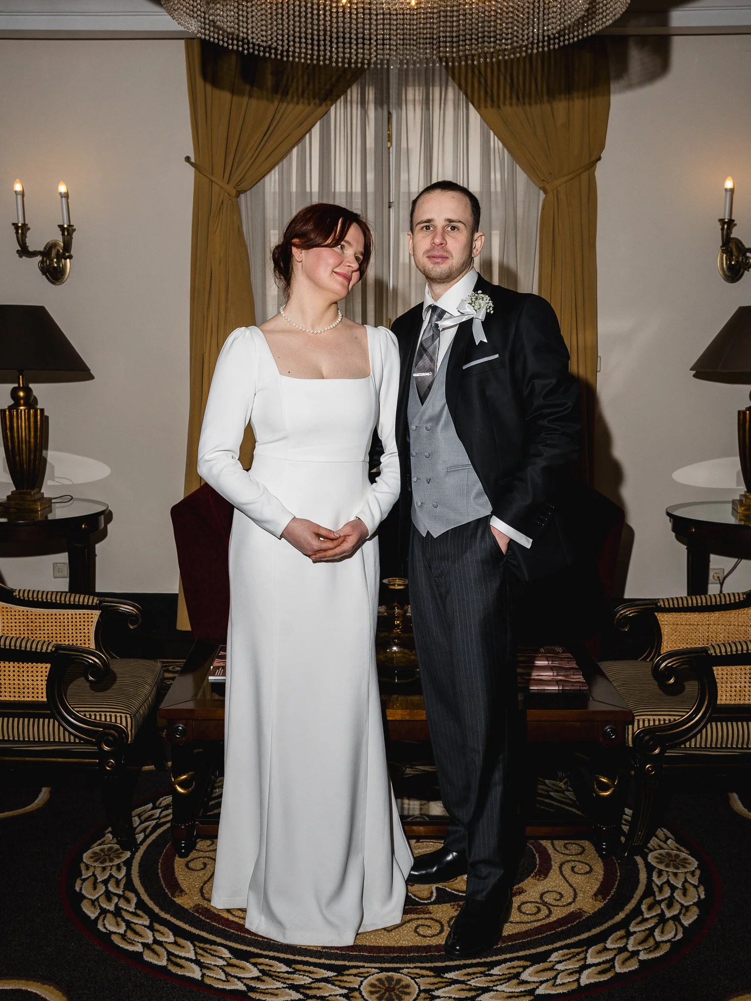 A bride and groom posing in a formal indoor setting, with the bride in a white wedding dress and the groom in a black tuxedo with a gray vest, standing under a chandelier, flanked by lamps and gold curtains.