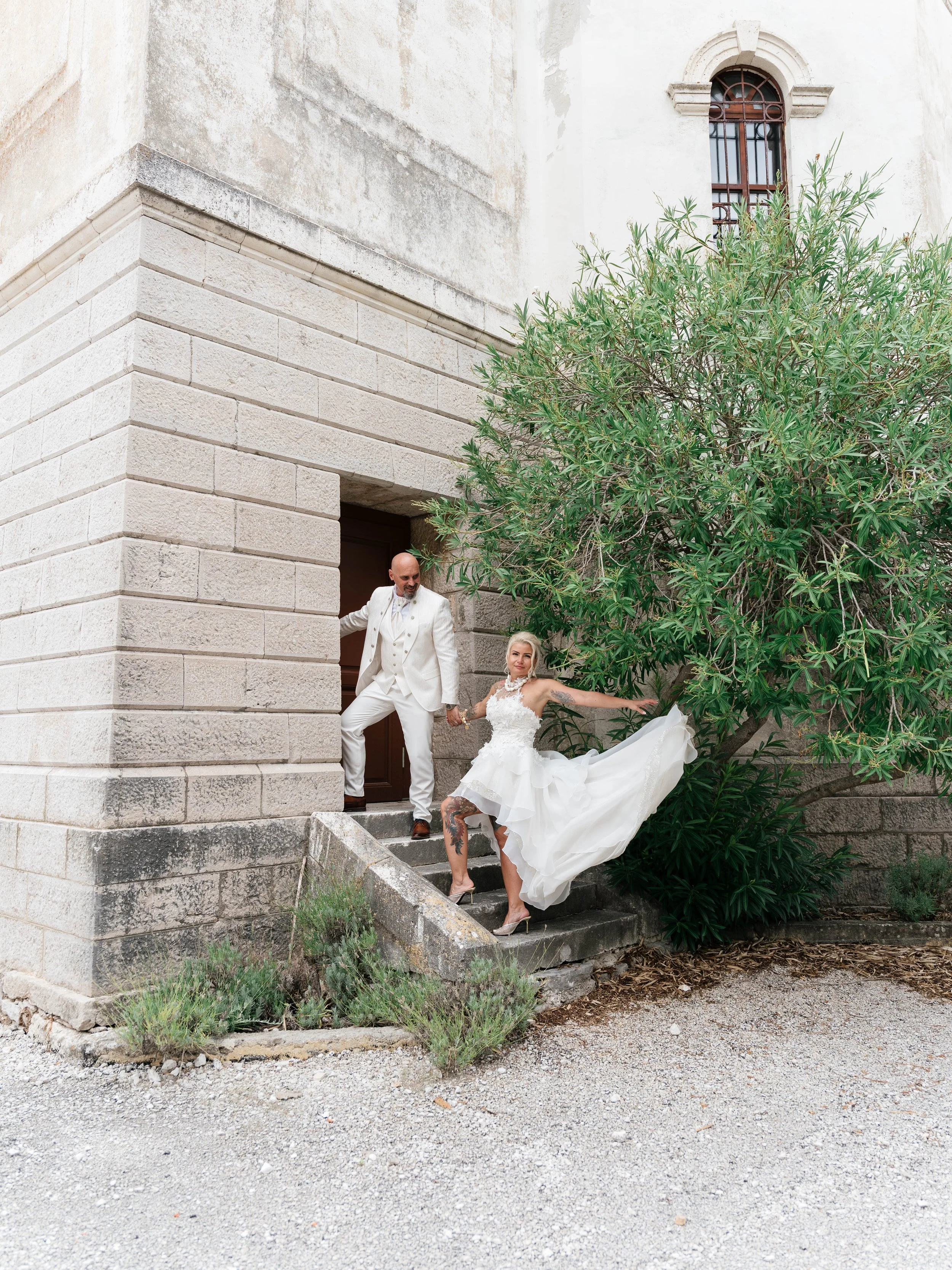 A couple dressed in wedding attire standing on a stone staircase outside a building with a large tree partially obscuring the scene. The woman, with tattoos on her legs, is in a white wedding dress and high heels, while the man, bald, is in a white s