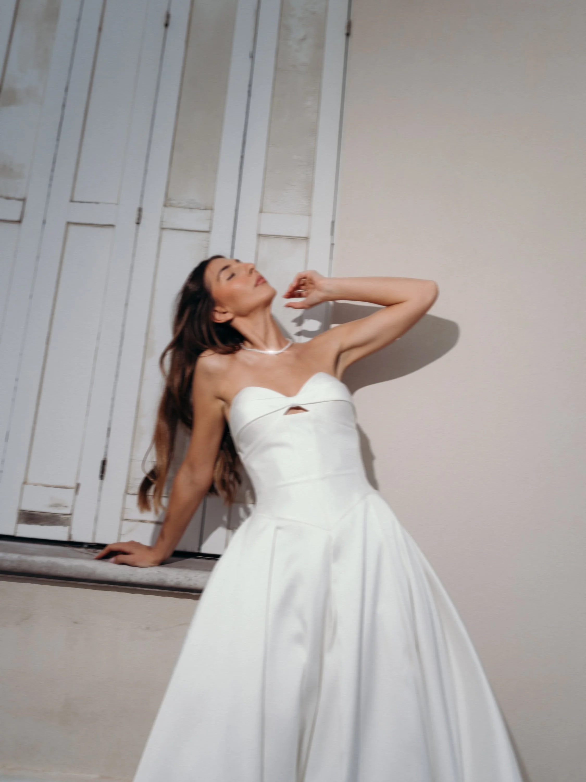 Woman in a white strapless dress posing with her head tilted back and one hand touching her face, standing by a white wall with closed window shutters.