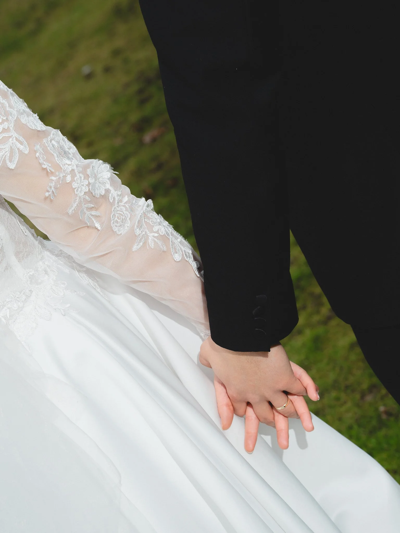 Close-up of a bride and groom holding hands, the bride's dress features lace sleeves, and the groom wears a black suit, outdoor setting with grass.