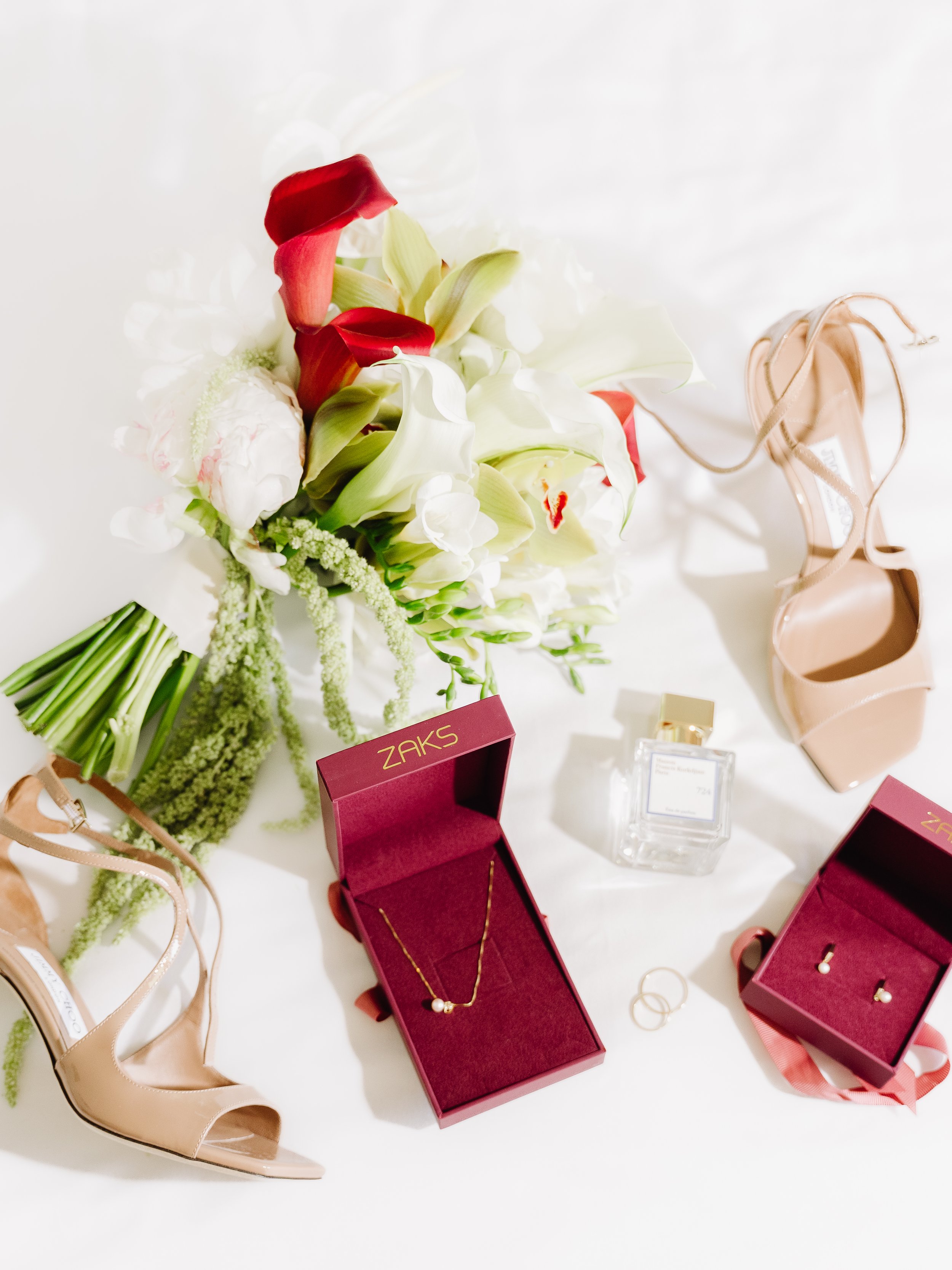 A display of a bridal bouquet with white and red flowers, several pairs of nude-colored high heel shoes, gold jewelry, and a jewelry box with a necklace and earrings inside, on a white surface.