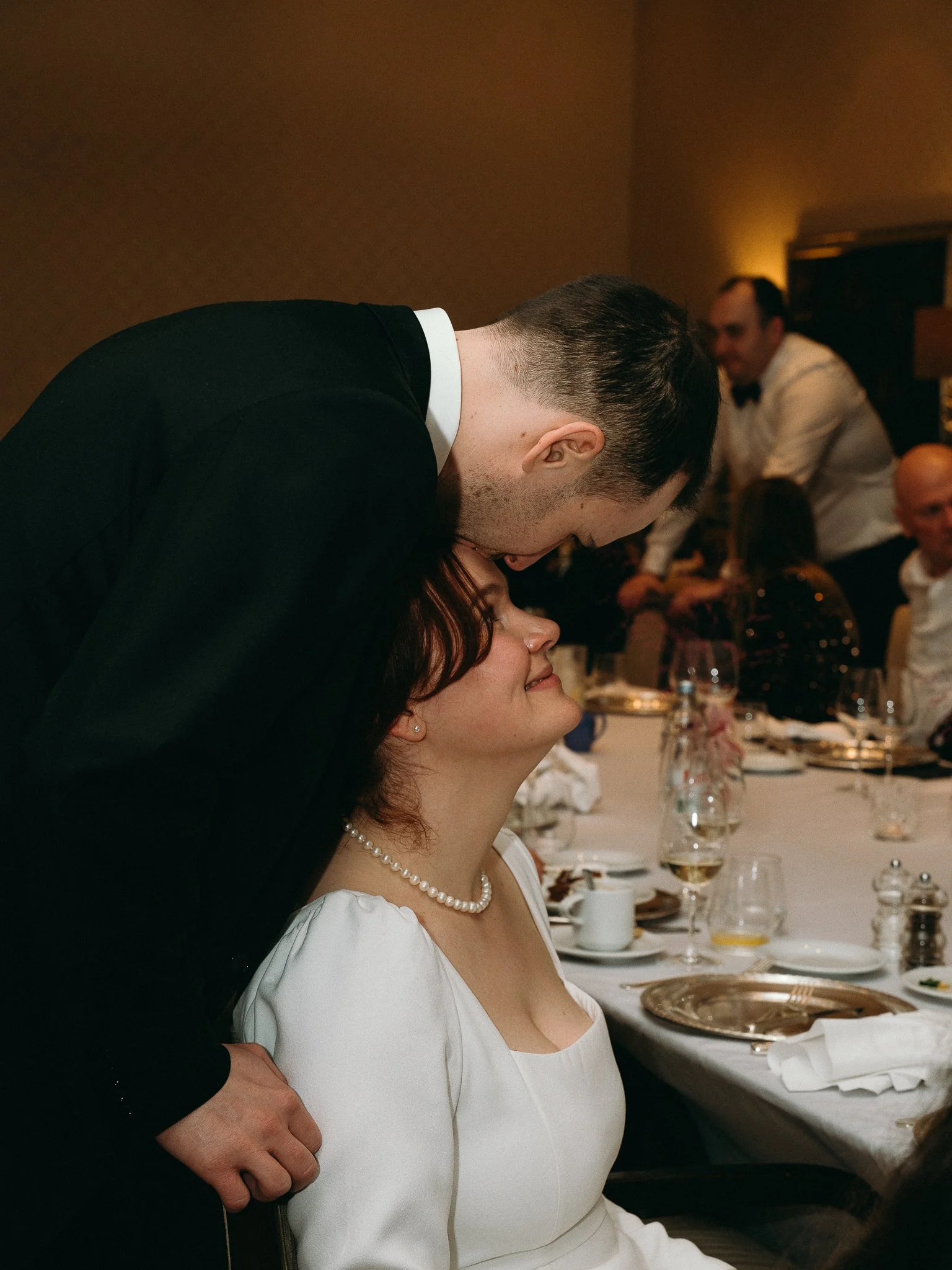 A man in a black suit and a woman in a white dress sharing a tender moment at a formal event, with the man leaning over and the woman smiling with closed eyes, sitting at a decorated table.