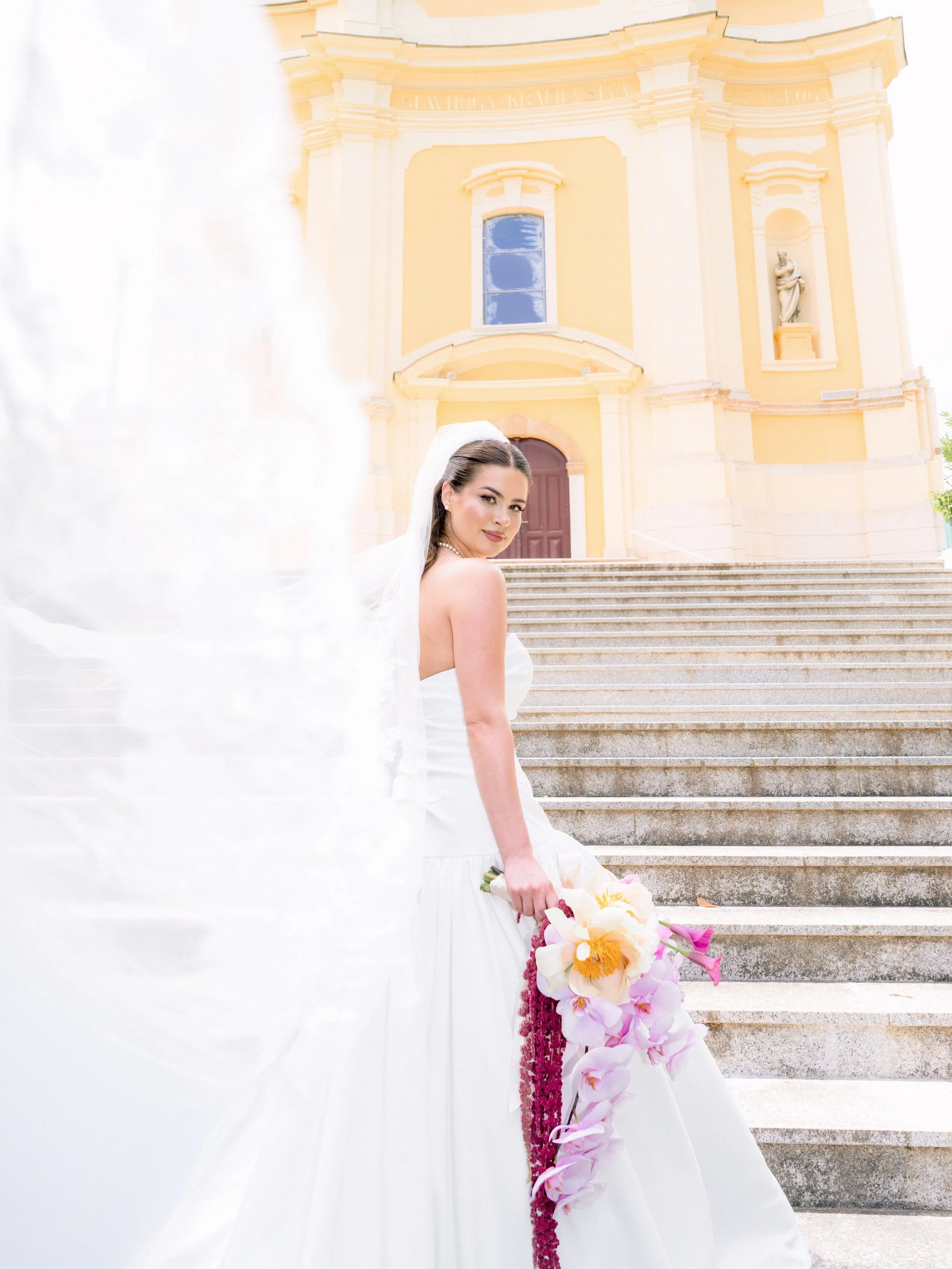 Loose editorial shot of a bride standing on stairs in front of a yellow church with a statue in a niche. She is holding a bouquet of pink and white flowers and wearing a white wedding dress with a veil.