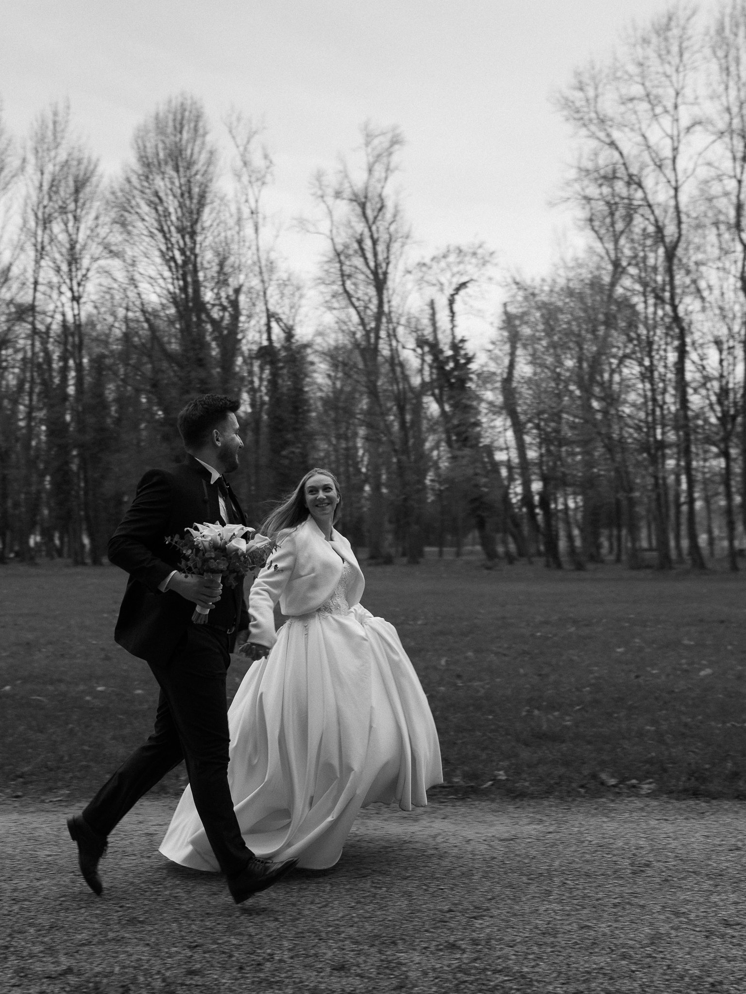 Black and white photo of a couple, the woman in a wedding dress and the man in a suit, holding hands and running outdoors in a park with trees in the background.