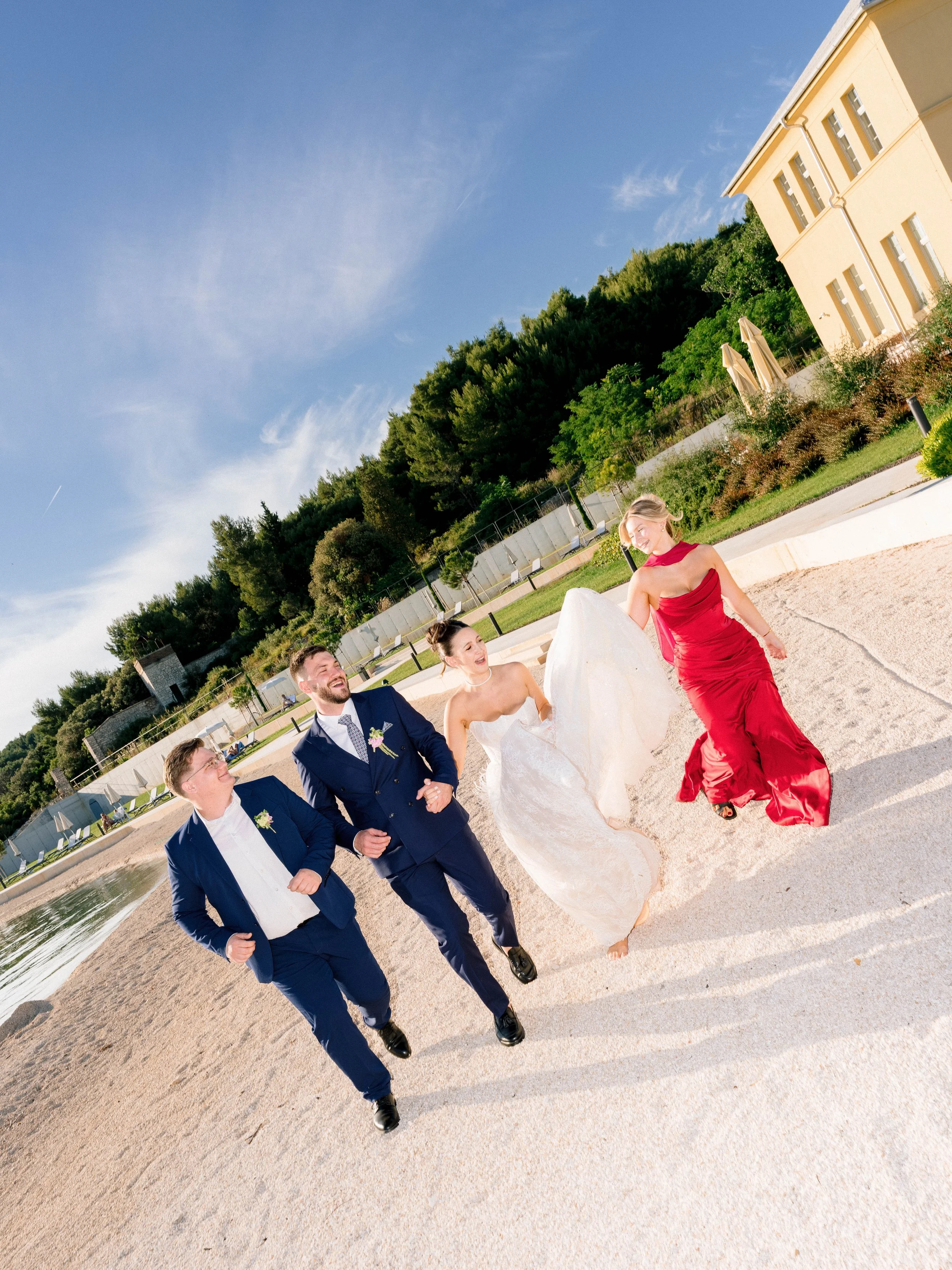 A group of five people walking on a sandy beach, including a woman in a red dress, a bride in a white gown, and three men in suits, with greenery and a yellow building in the background, under a blue sky.