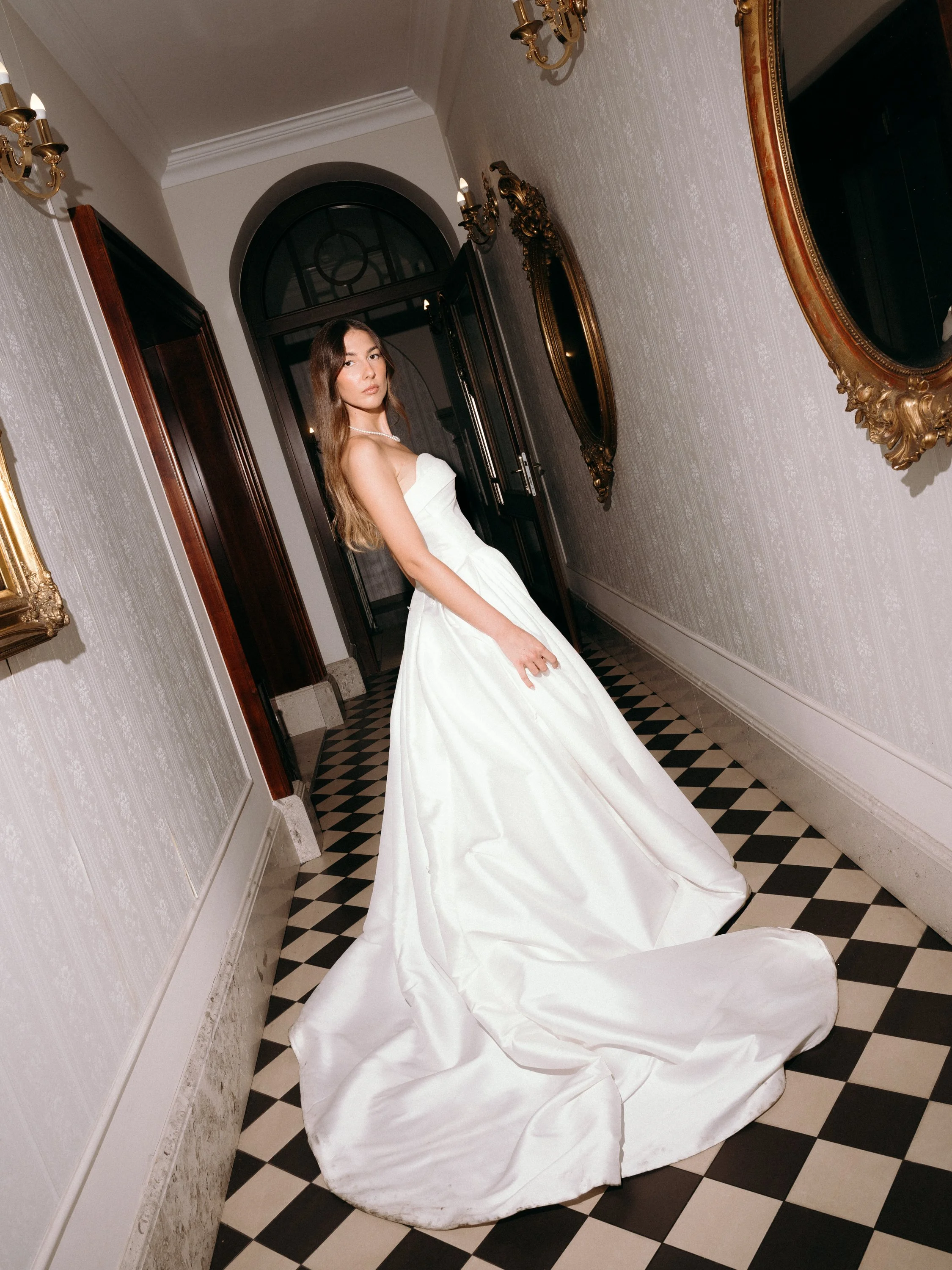 A young woman in a strapless white wedding gown standing in a hallway with ornate gold-framed mirrors and vintage wall sconces, on a black and white checkered floor.