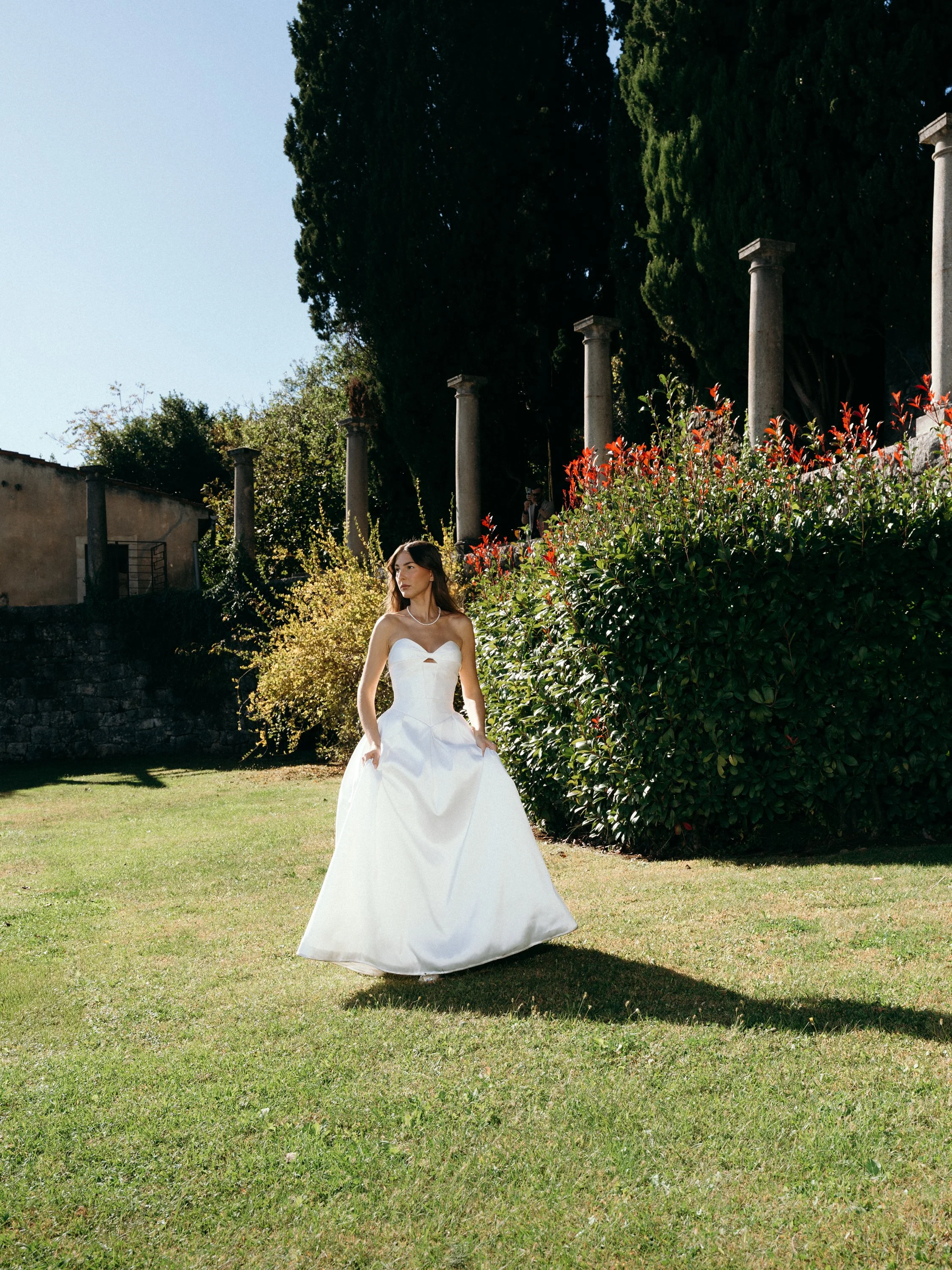 A woman in a white wedding dress standing on a grassy lawn near bushes and tall trees with columns in the background.